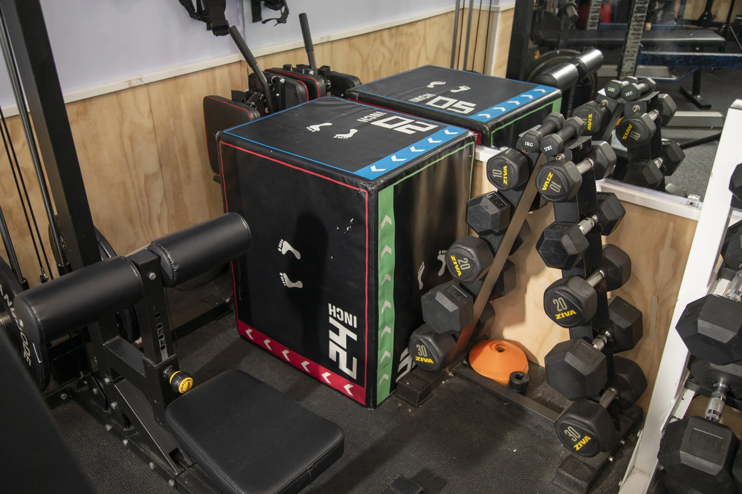 A gym corner with a plyometric box, adjustable dumbbells, and a mirror. The plyometric box has markings for different heights and weight categories. The dumbbells are arranged on a rack, with weights labeled in kilograms. The mirror reflects part of 
