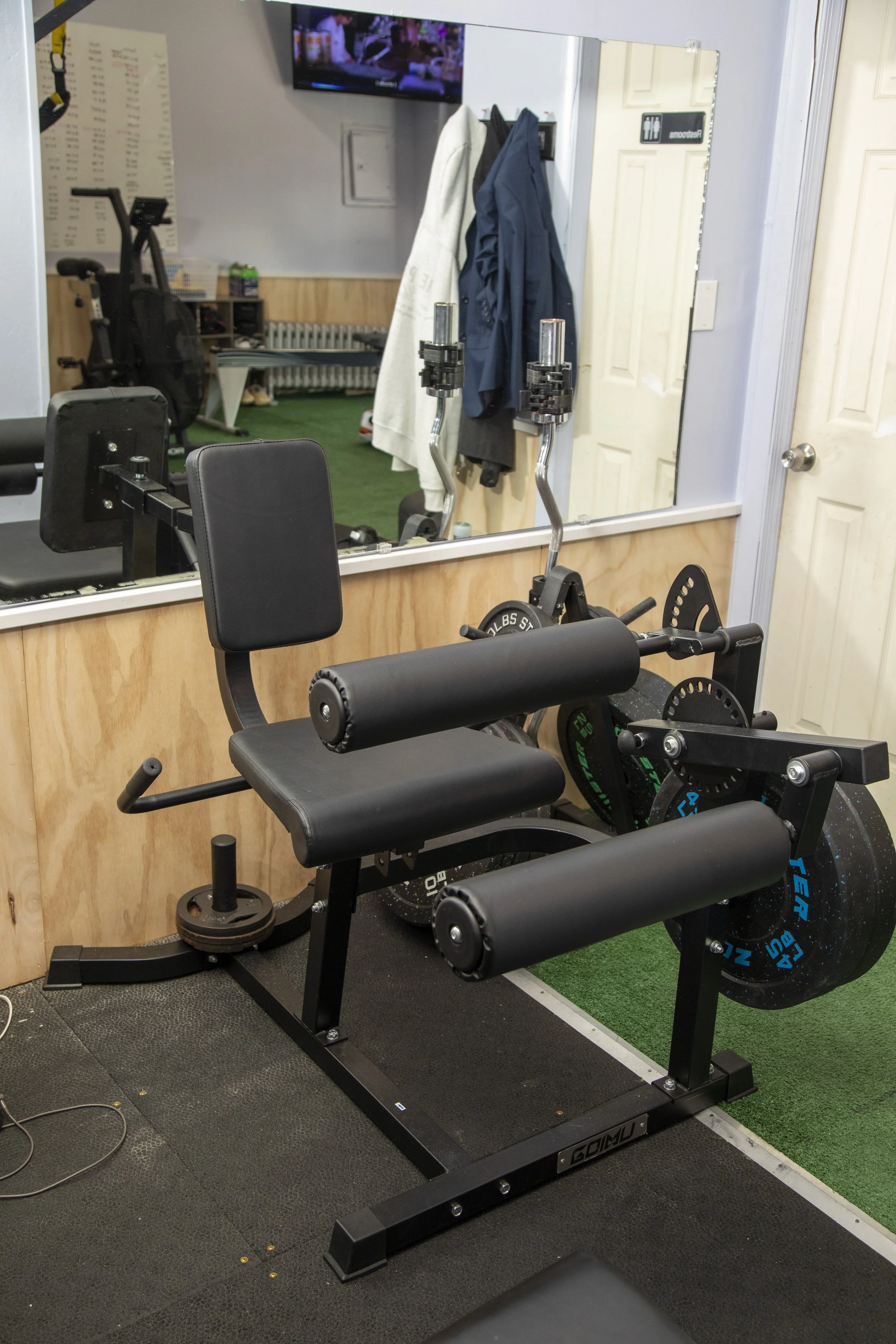 Gym equipment including leg curl machine, weight plates, and a mirror, in a fitness room with a TV, jackets hanging, a door with a restroom sign, and a green turf floor patch.
