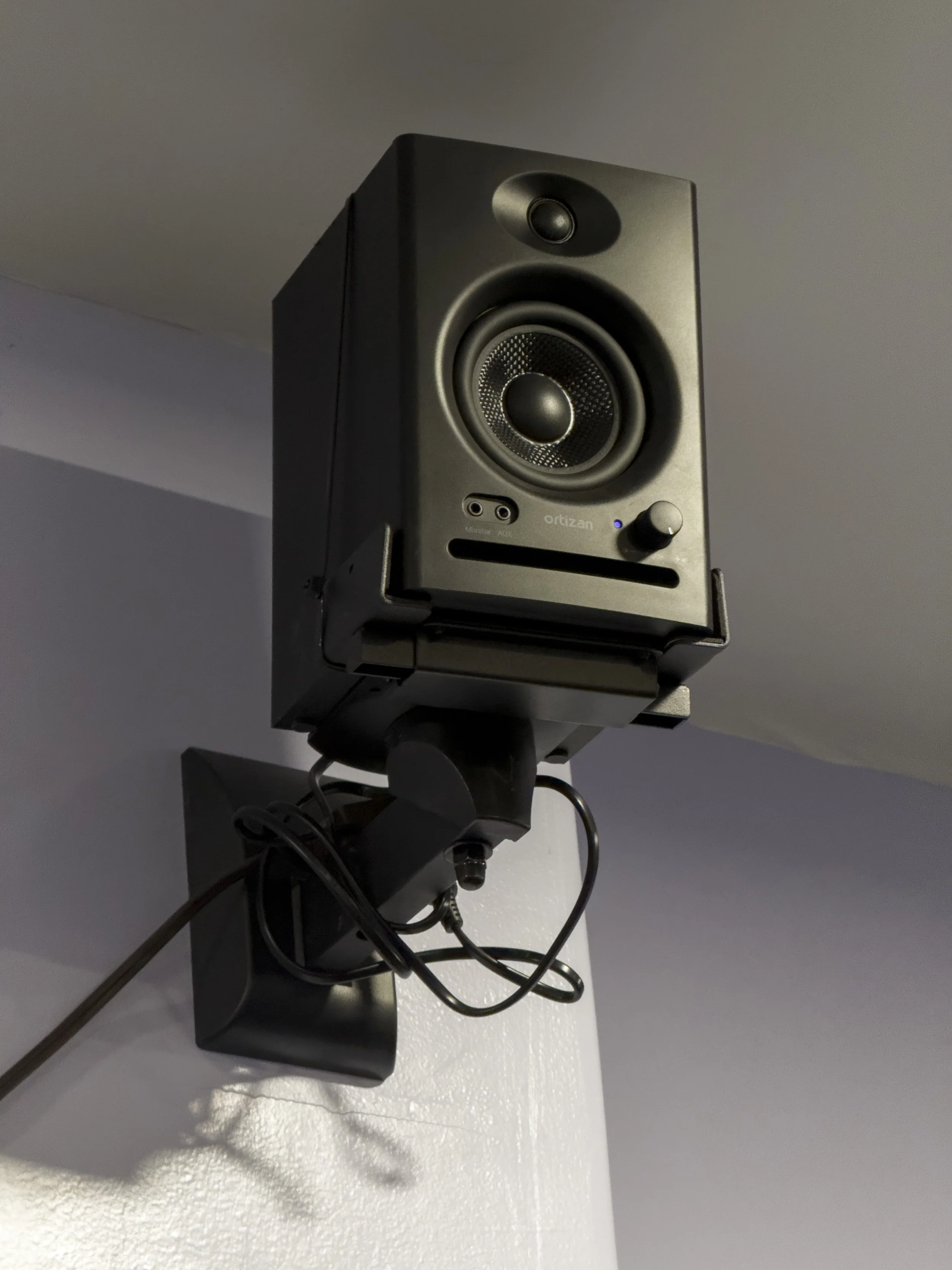 A black studio monitor speaker mounted on a white wall with visible cables attached.