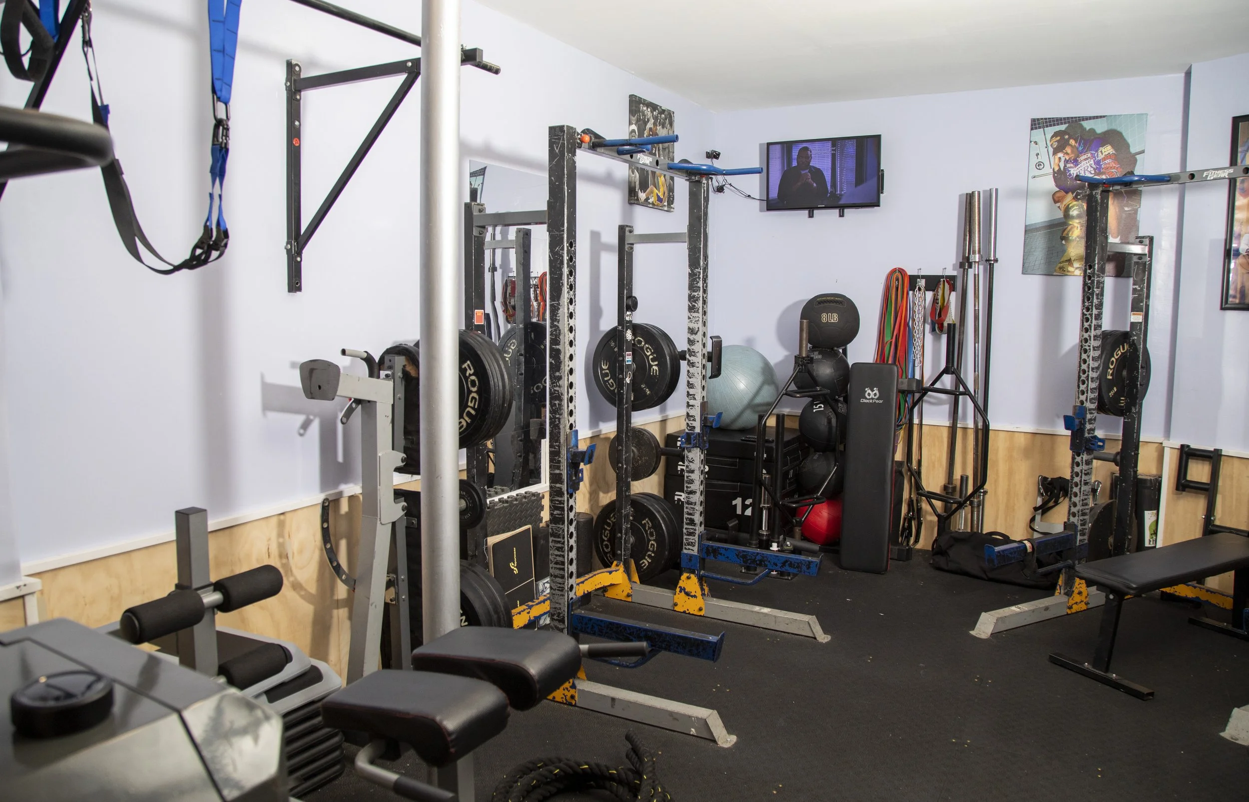 A small gym room with various workout equipment including weight plates, resistance bands, and a pull-up bar. There is a wall-mounted TV and posters in the background.