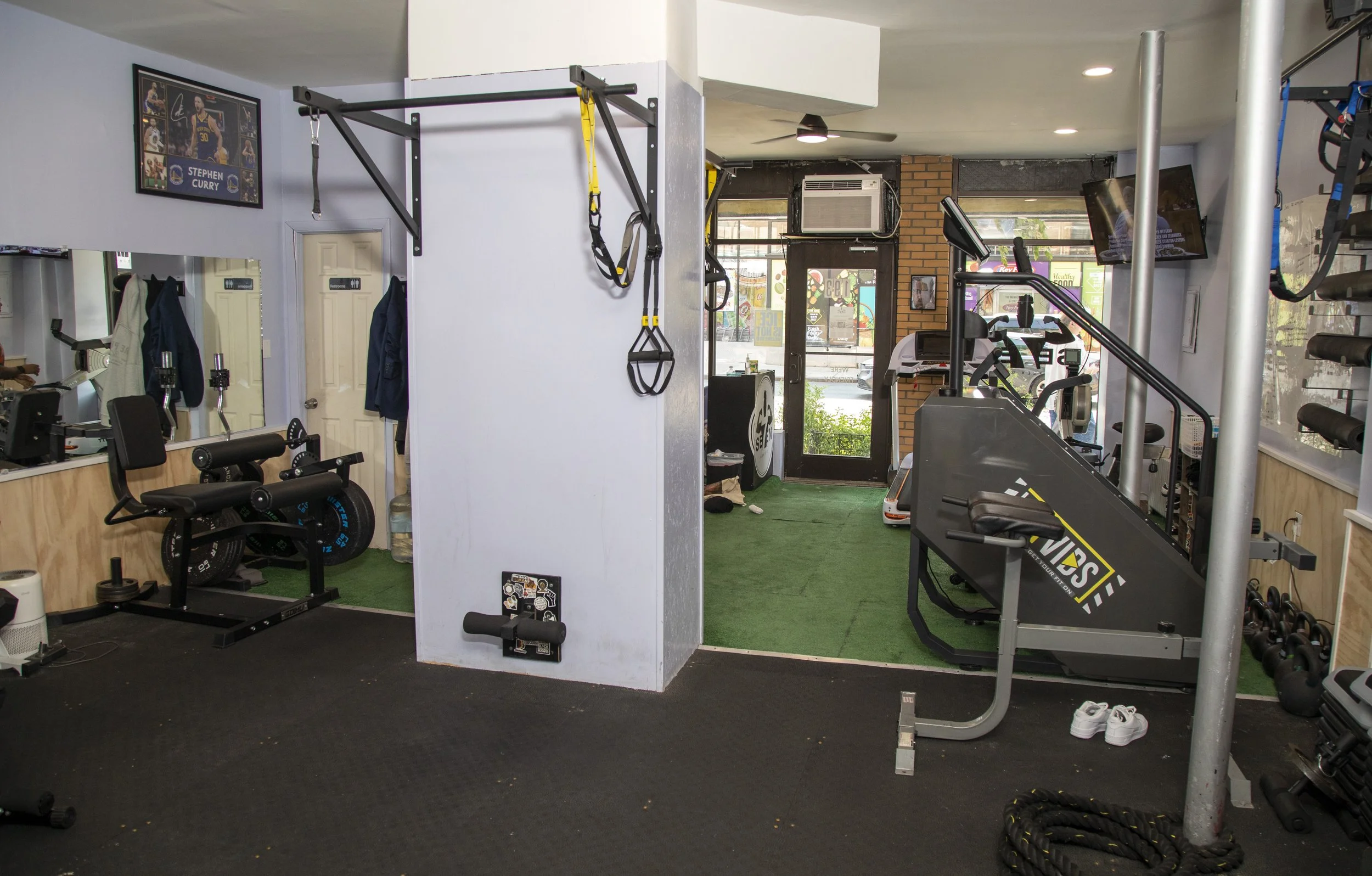 Interior of a gym with exercise equipment including a treadmill, rowing machine, resistance bands, and a pull-up station, with a wall-mounted TV and a framed poster of Stephen Curry.