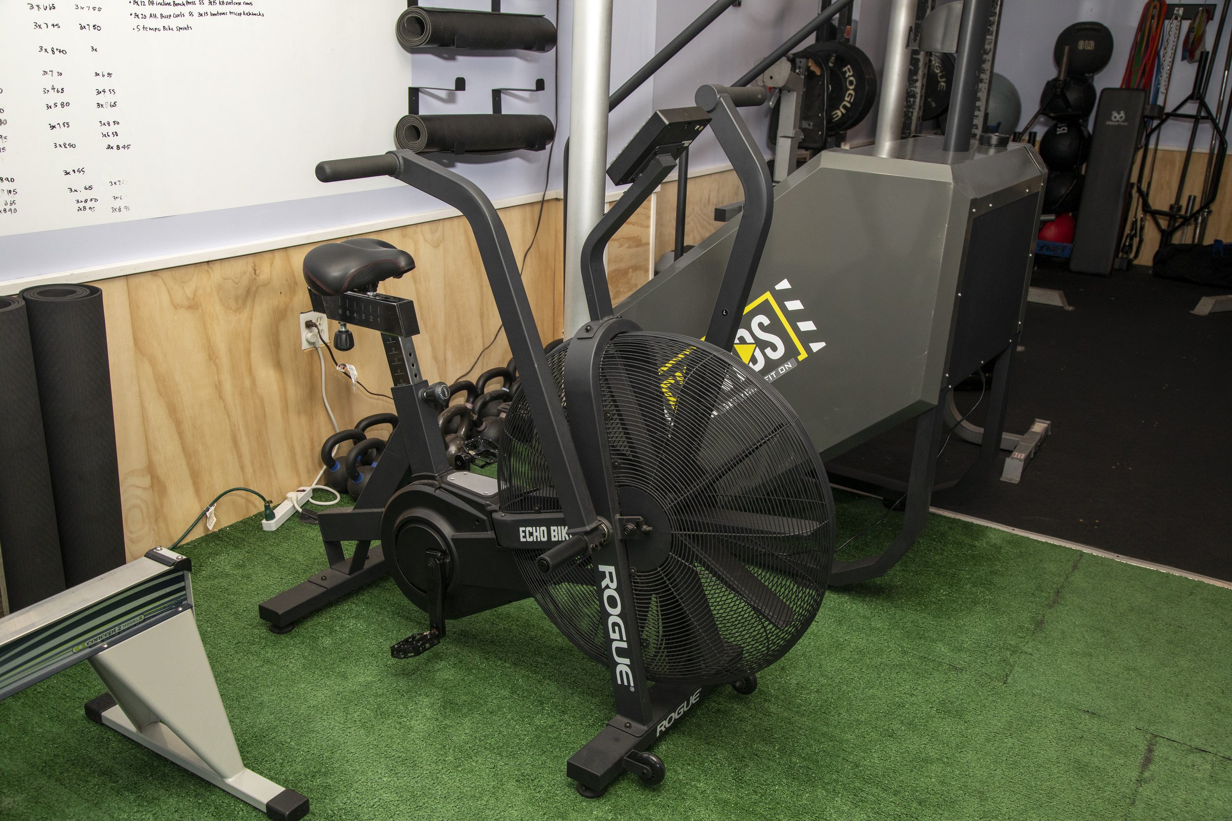 Indoor gym with exercise equipment including a rowing machine, stationary bike, and various weights, with a whiteboard and foam rollers on the wall.