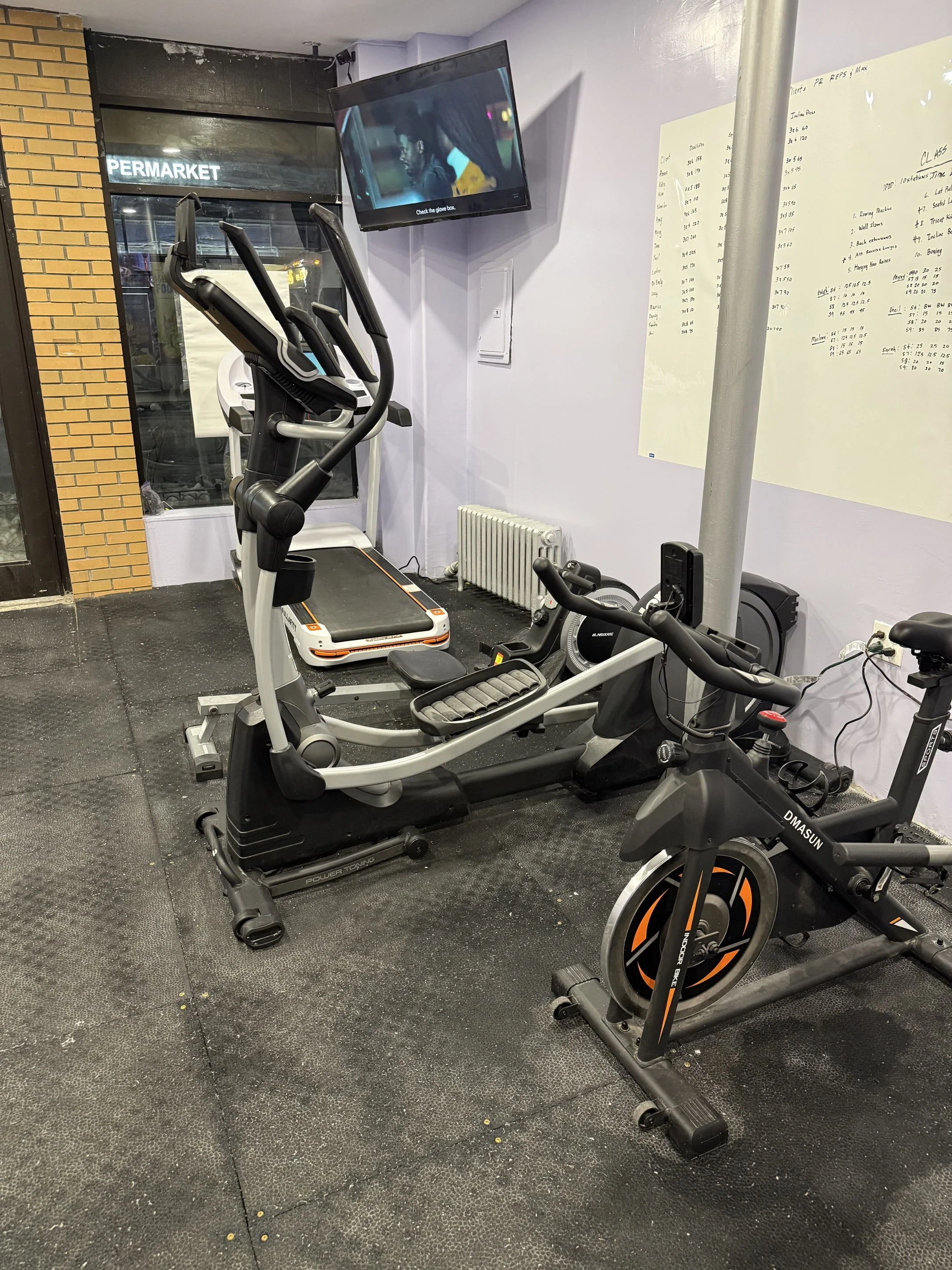 Indoor gym with an elliptical machine, stationary bike, and treadmill. A wall-mounted TV displays a man with a cigarette. Whiteboard with workout notes is on the wall, and a radiator is below it. Exterior view shows a sign for 'PERMARCKET'.