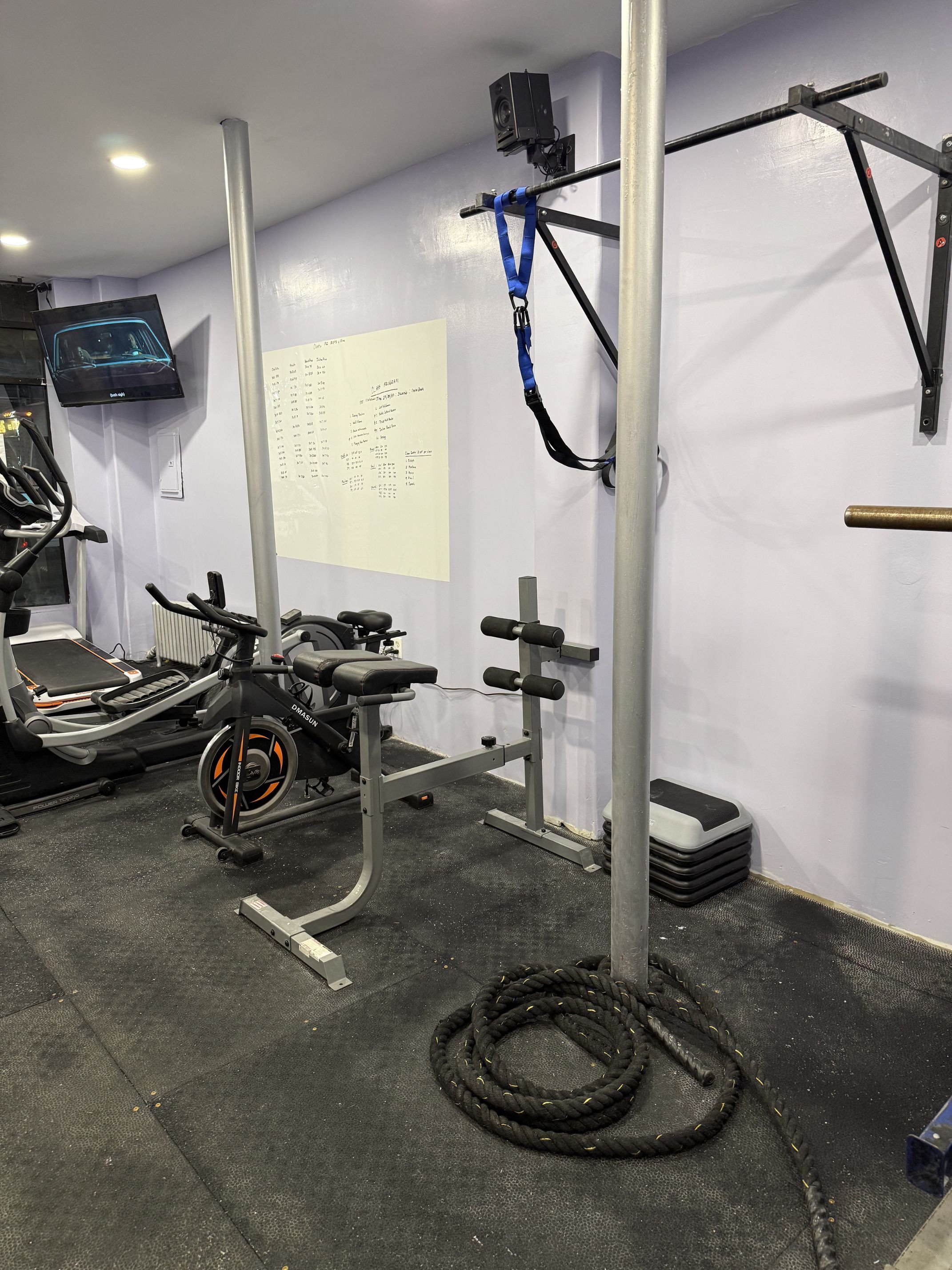 Gym equipment including a treadmill, stationery bike, a grip strength trainer, weight stack, TRX suspension trainer, coiled battle ropes, and a small stack of aerobic steps in a workout space.