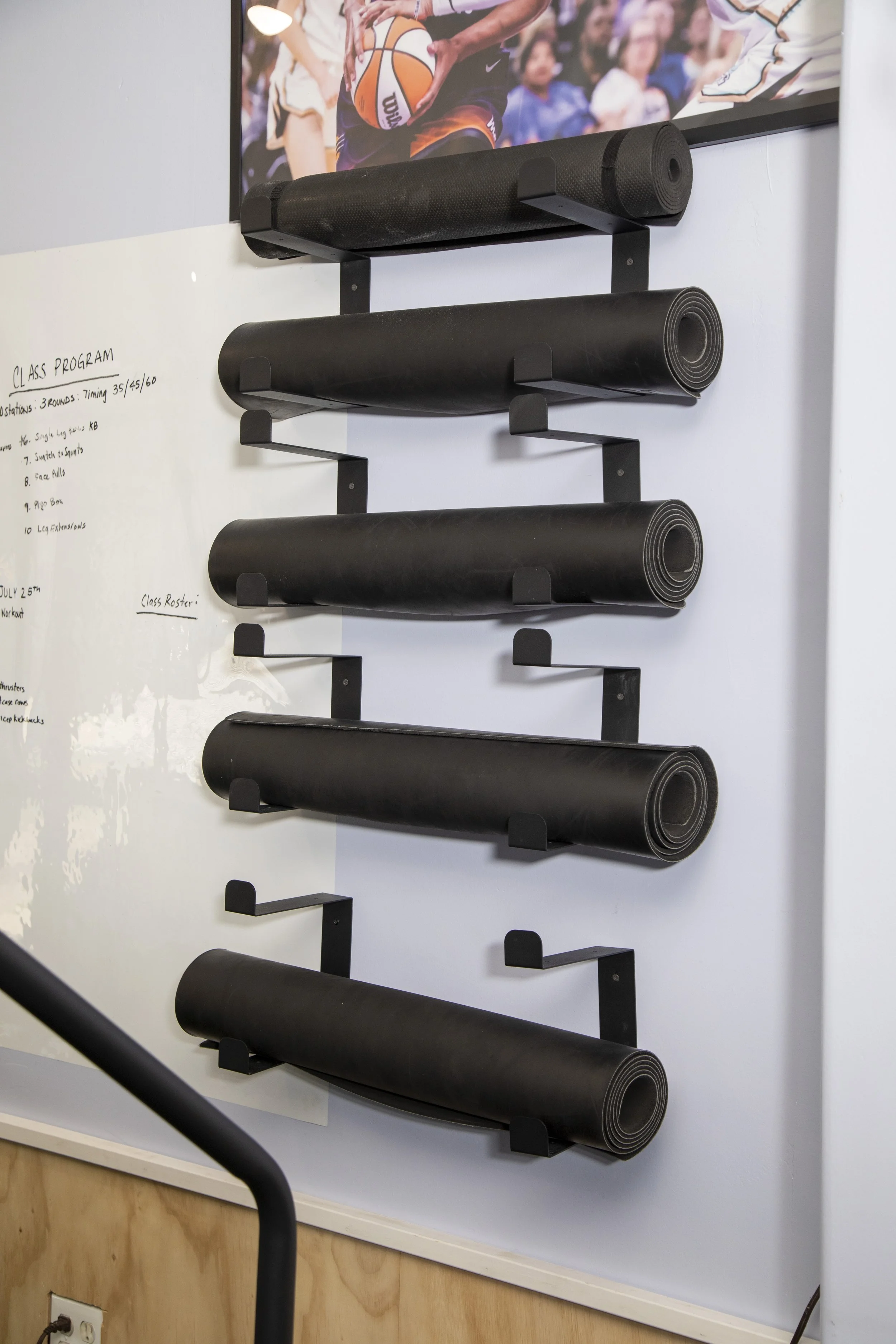 Wall-mounted black exercise mats in a gym, with a basketball game picture hanging above. Whiteboard with handwritten workout plan next to the mats, and a wooden panel below.