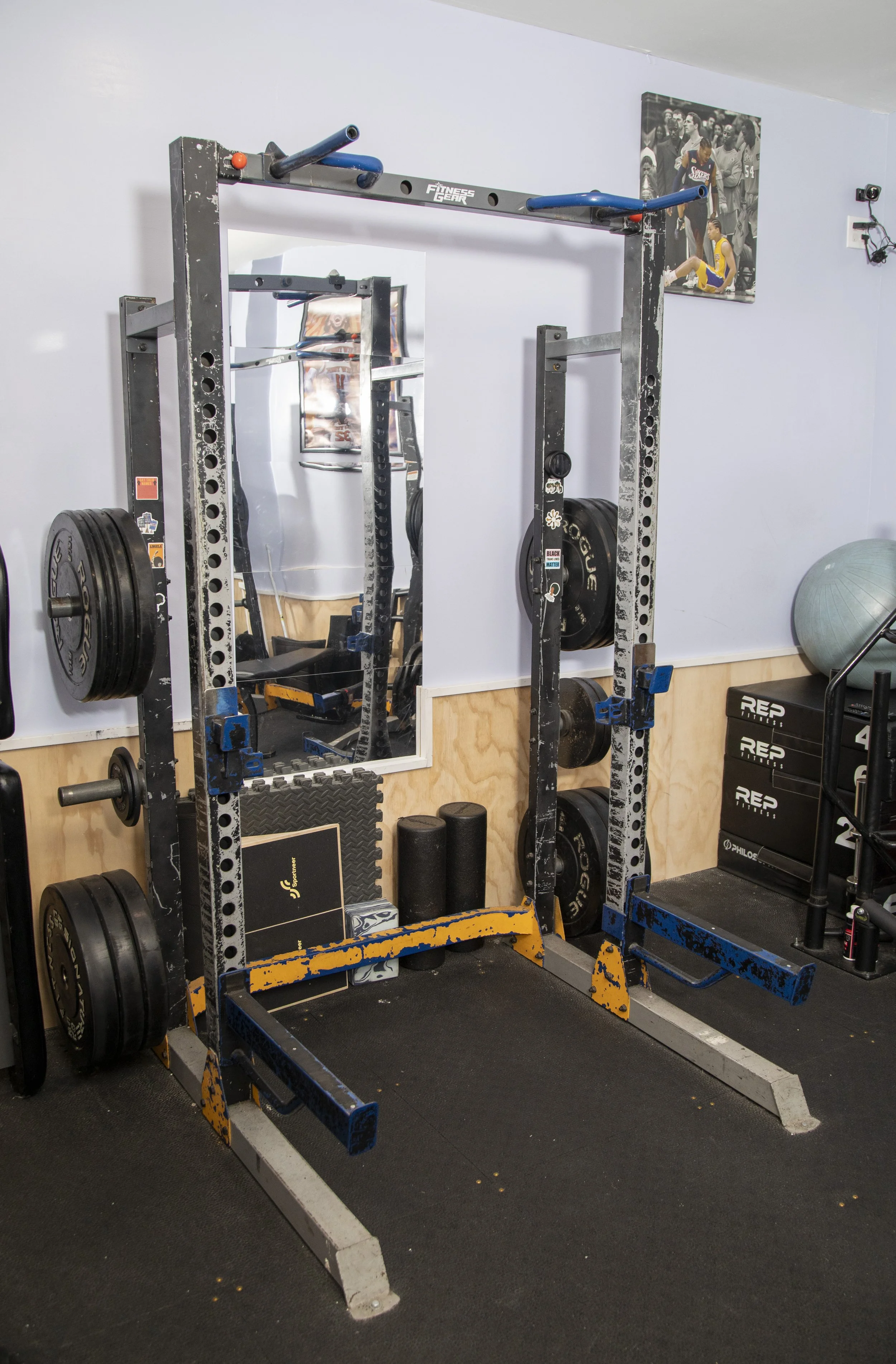 Indoor gym with weightlifting equipment, including a squat rack with weight plates, a mirror, a black foam roller, a stability ball, and fitness posters on the wall.
