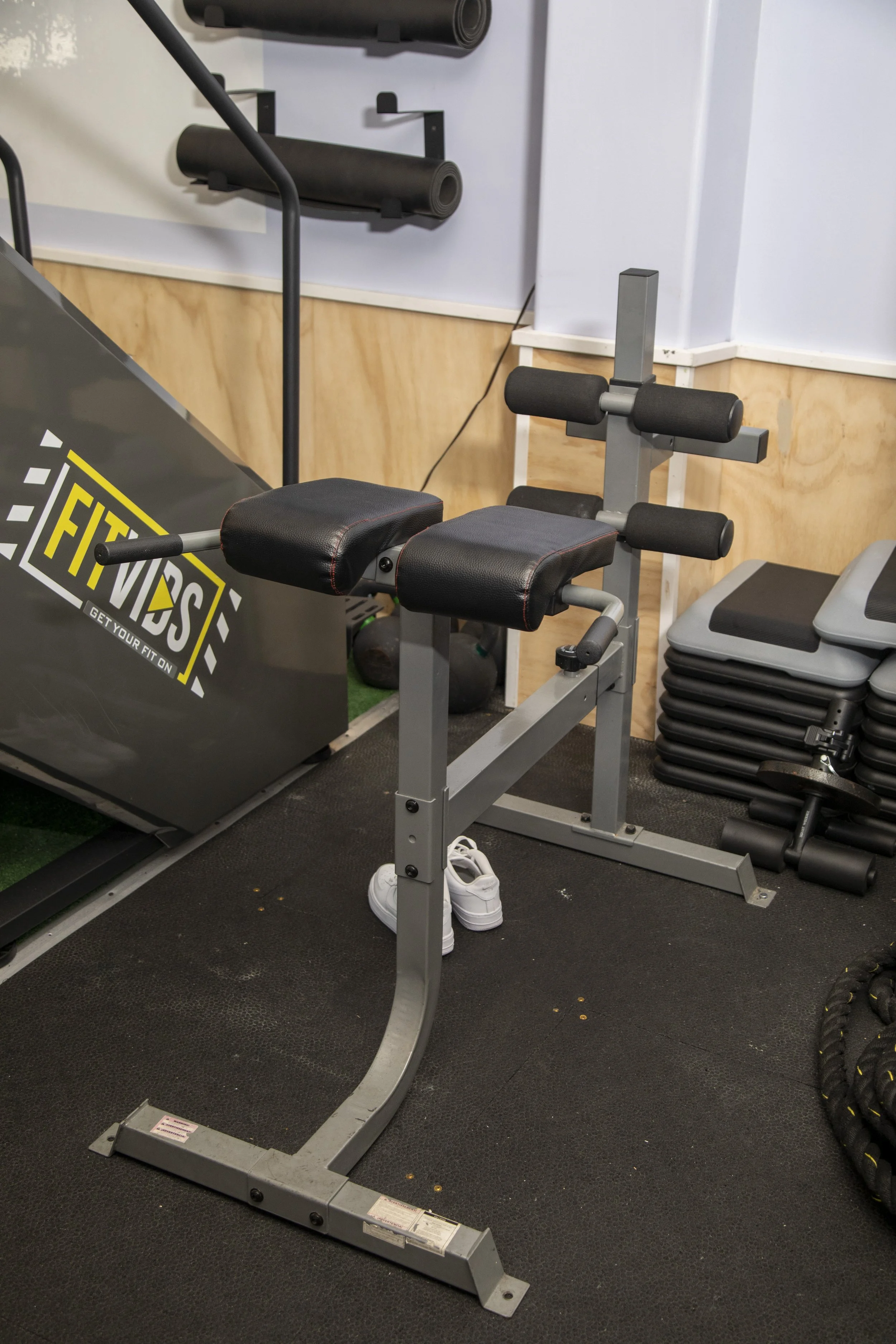 A gym space featuring a rowing machine with black padded support, a stack of black aerobic steps, a weightlifting machine with foam rollers, and wall-mounted black rollers for stretching or exercise.