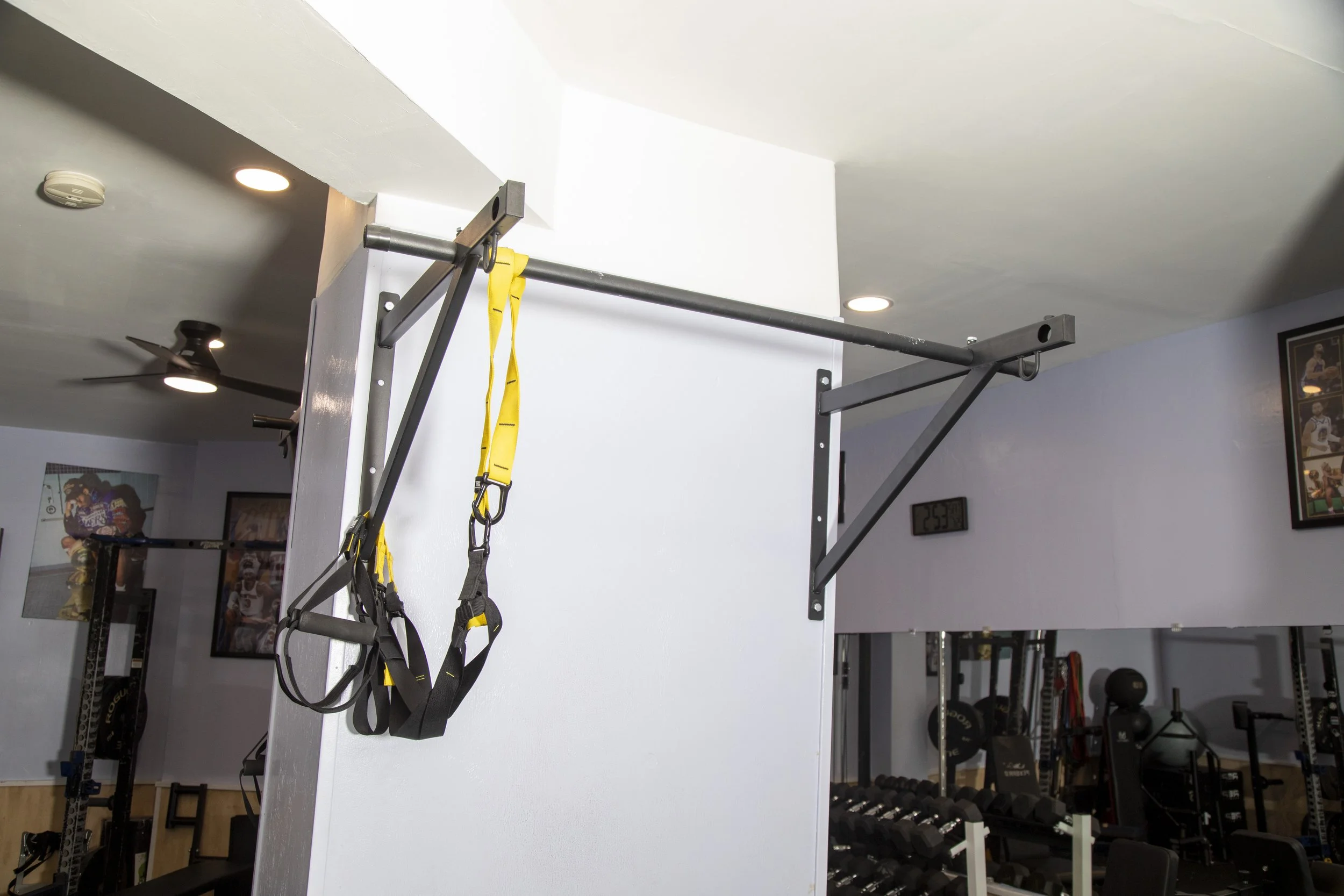 Gym area with TRX suspension straps hanging from a metal frame attached to the ceiling, with a white wall, a thermostat, and exercise equipment including dumbbells in the background.