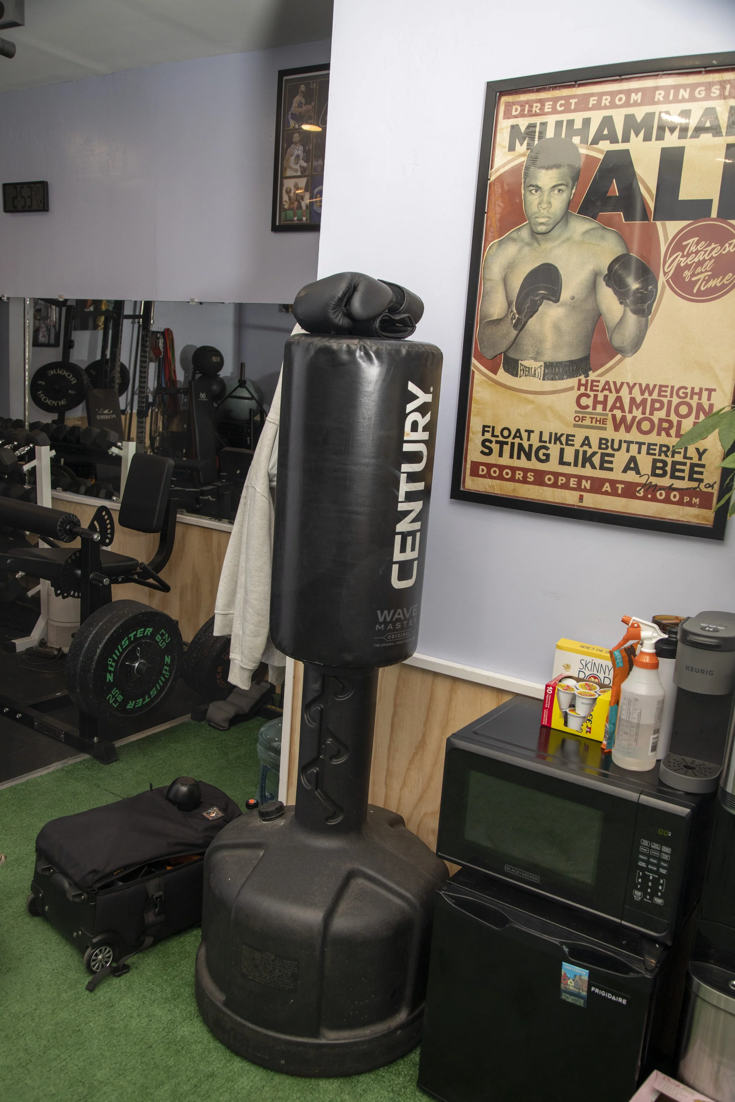 Indoor gym space with boxing equipment including a punching bag, padded gloves, and various weights. The wall features a vintage boxing poster of Muhammad Ali, and there are workout accessories like a microwave, snacks, and cleaning supplies on a cou