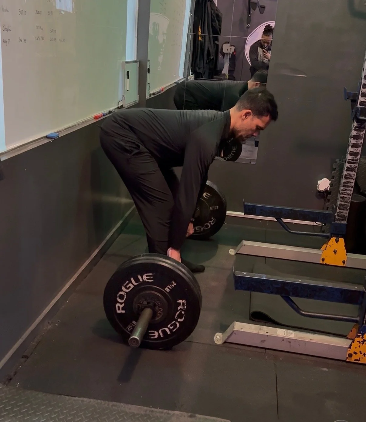 A man wearing black workout clothes bending over to pick up a barbell with weight plates, inside a gym with black walls and a whiteboard, with a person taking a photo in the mirror.