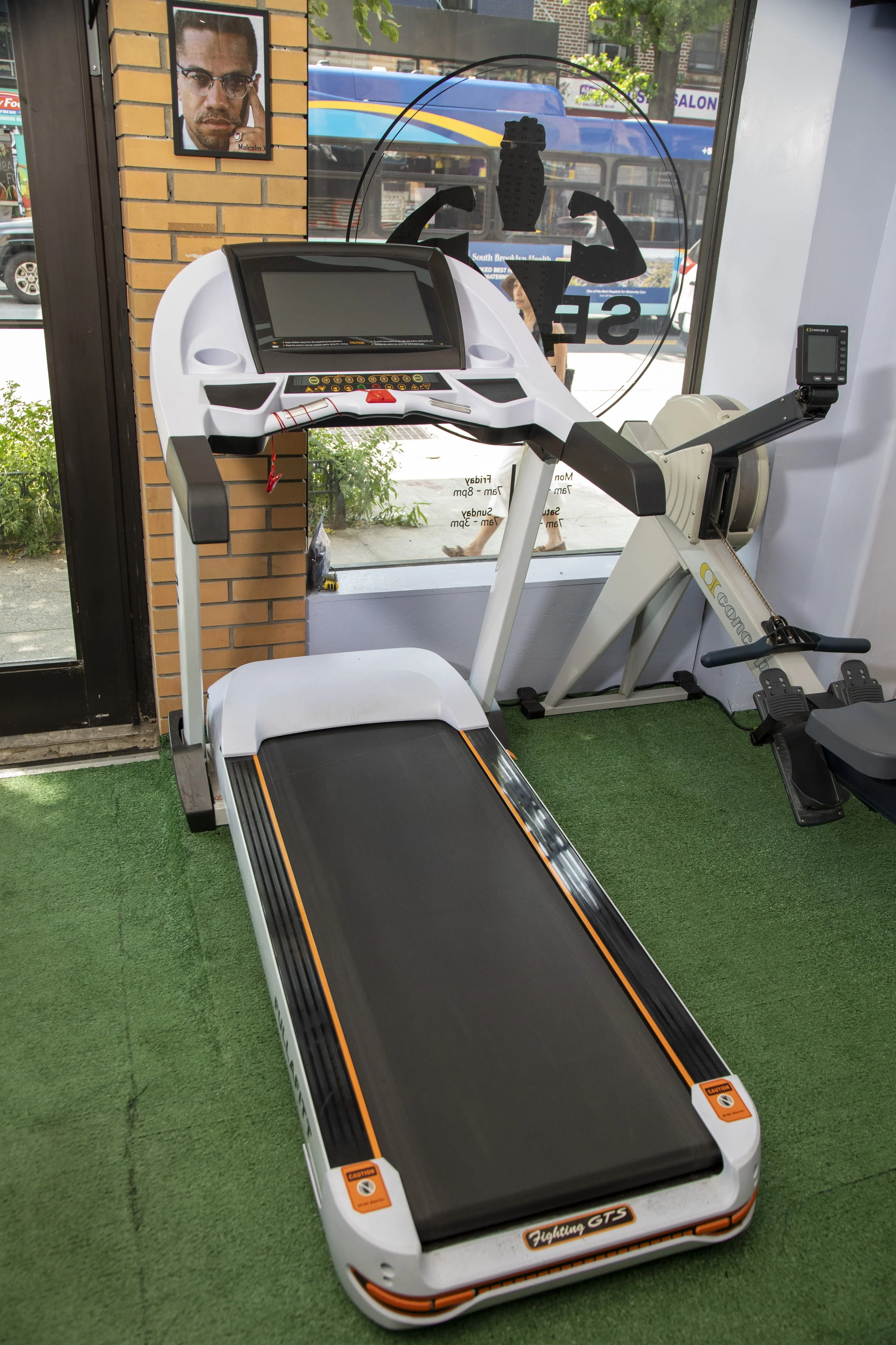 Indoor gym equipment, including a treadmill and an elliptical machine with a large digital screen, next to a window with a view of a street with a bus, pedestrians, and a brick wall with a portrait poster.