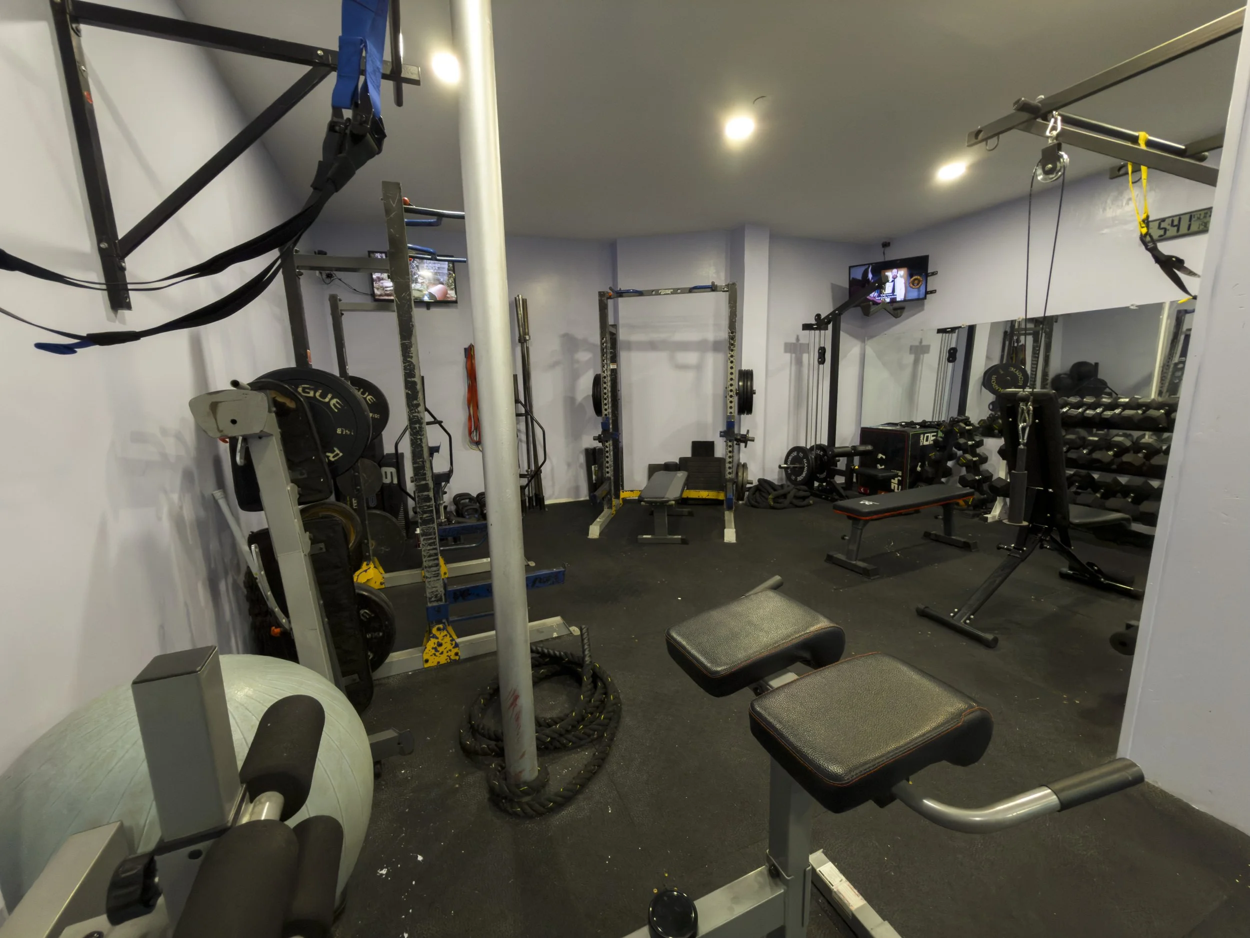 A small gym with various workout equipment including dumbbells, weight plates, a bench, a rowing machine, pull-up bars, resistance bands, a stability ball, and televisions mounted on the walls.