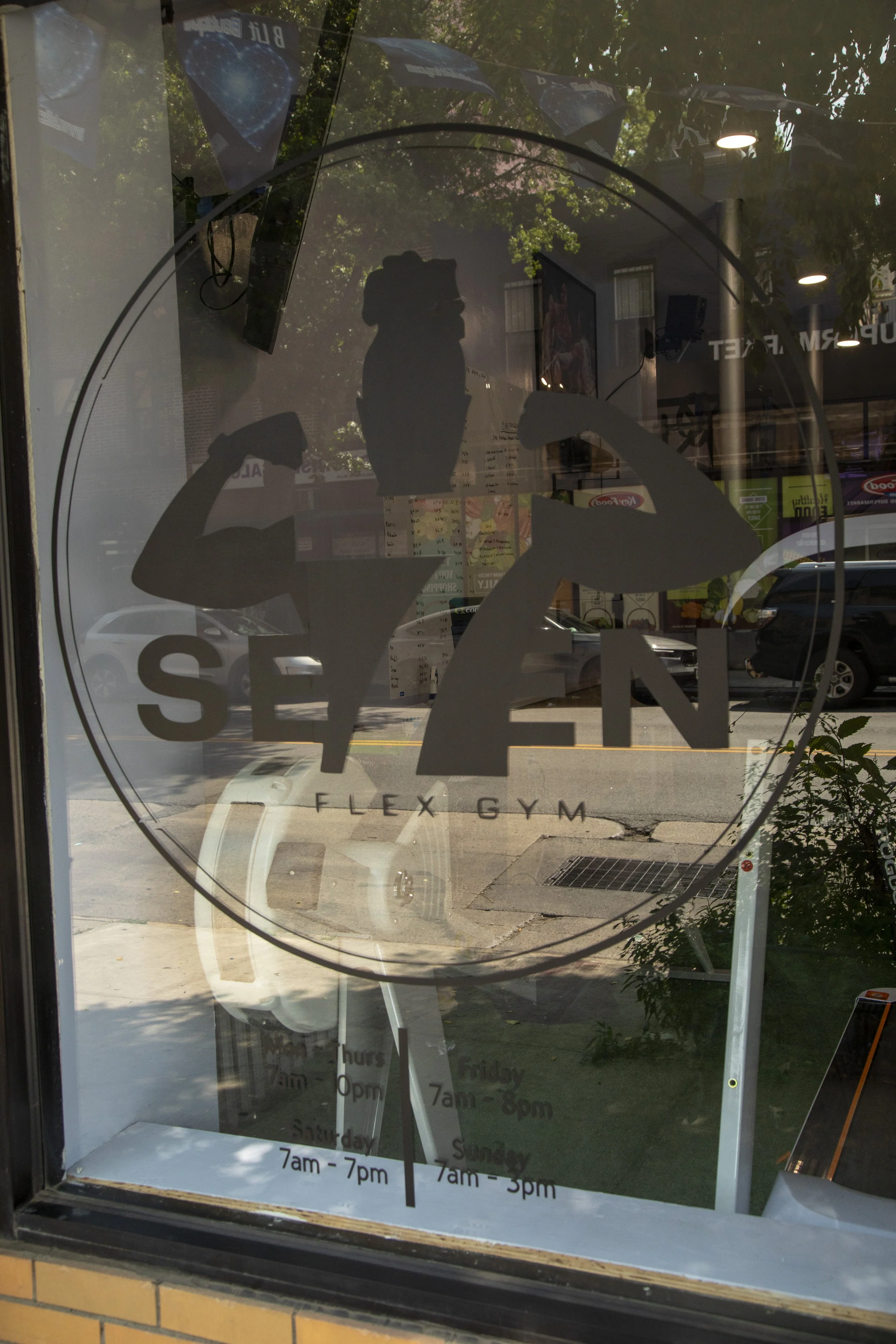 Reflection of a person taking a photo is visible on a glass window of a gym, with trees and parked cars outside. The window displays a large logo of a flexed bicep muscle and text indicating gym hours.