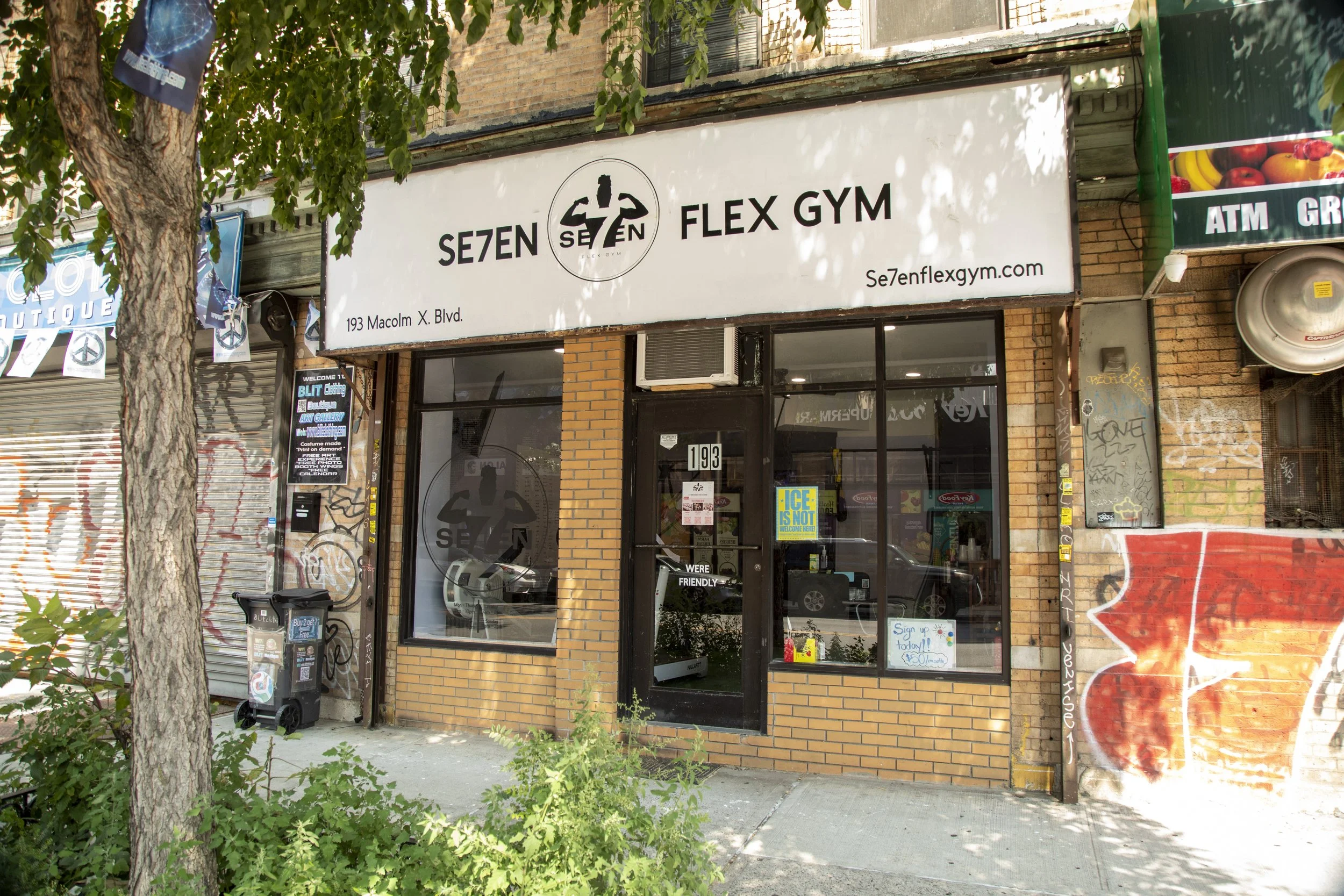  storefront of Se7en Flex Gym with brick exterior, large windows, and a white sign showing the gym's name, address 193 Macolm X. Blvd., and website se7enflexgym.com. Some stickers and graffiti are visible on the walls and window. There are trees and 