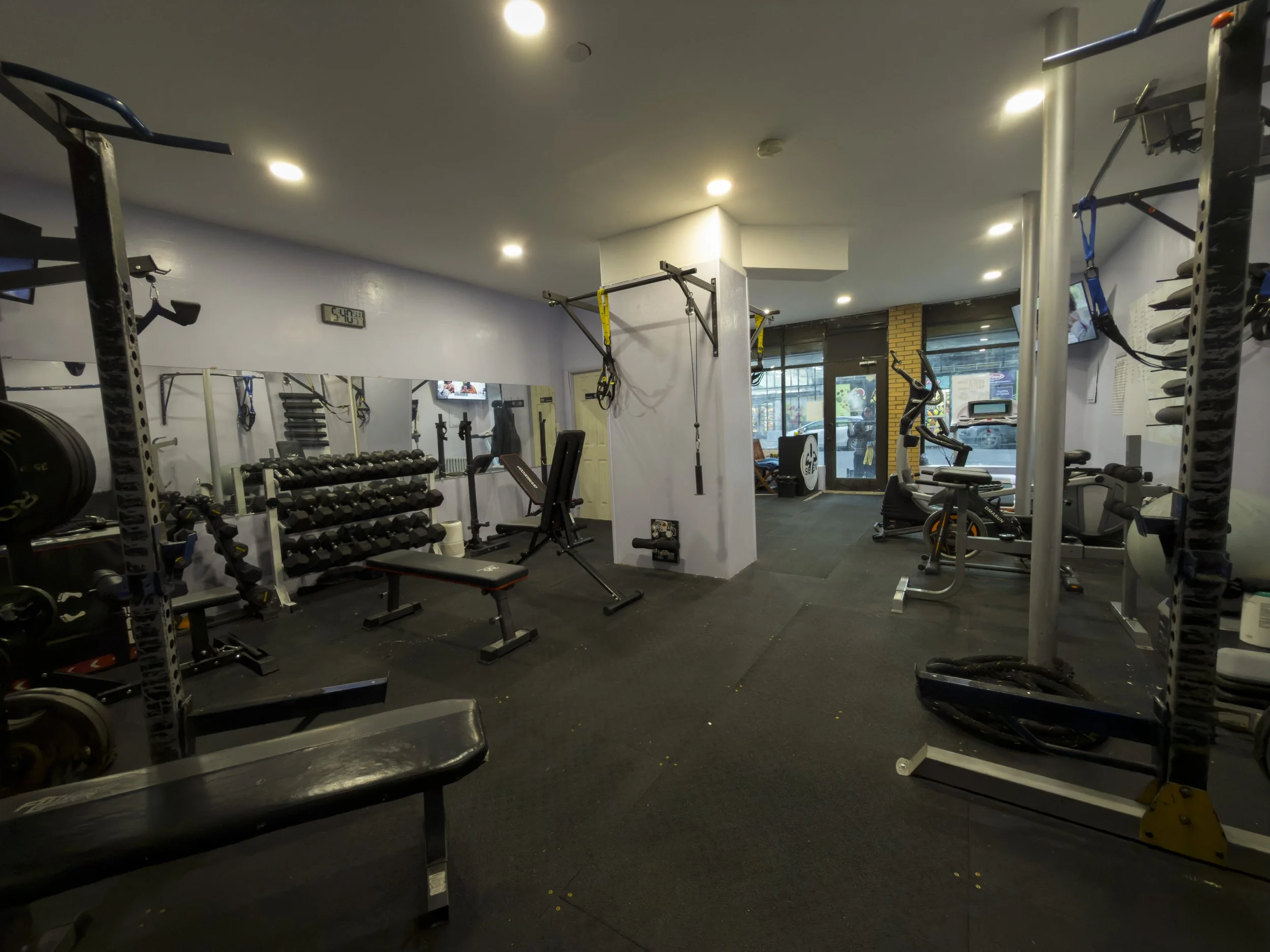 A gym with various exercise equipment including weights, machines, and cardio equipment, with mirrors on the wall and a window showing outside view.