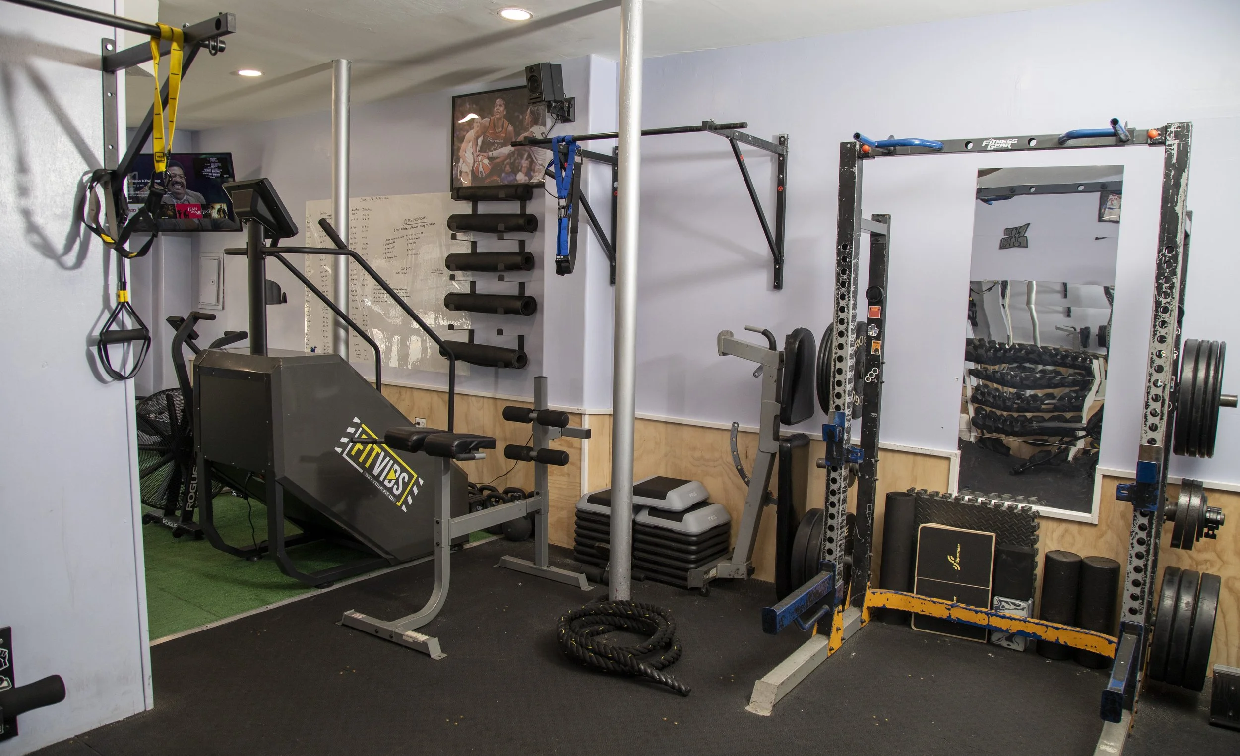 Interior of a small gym with various workout equipment including a treadmill, pull-up bars, weight racks, and medicine balls. There are two TV screens mounted on the wall showing athletes, and the floor is covered with black rubber mats.