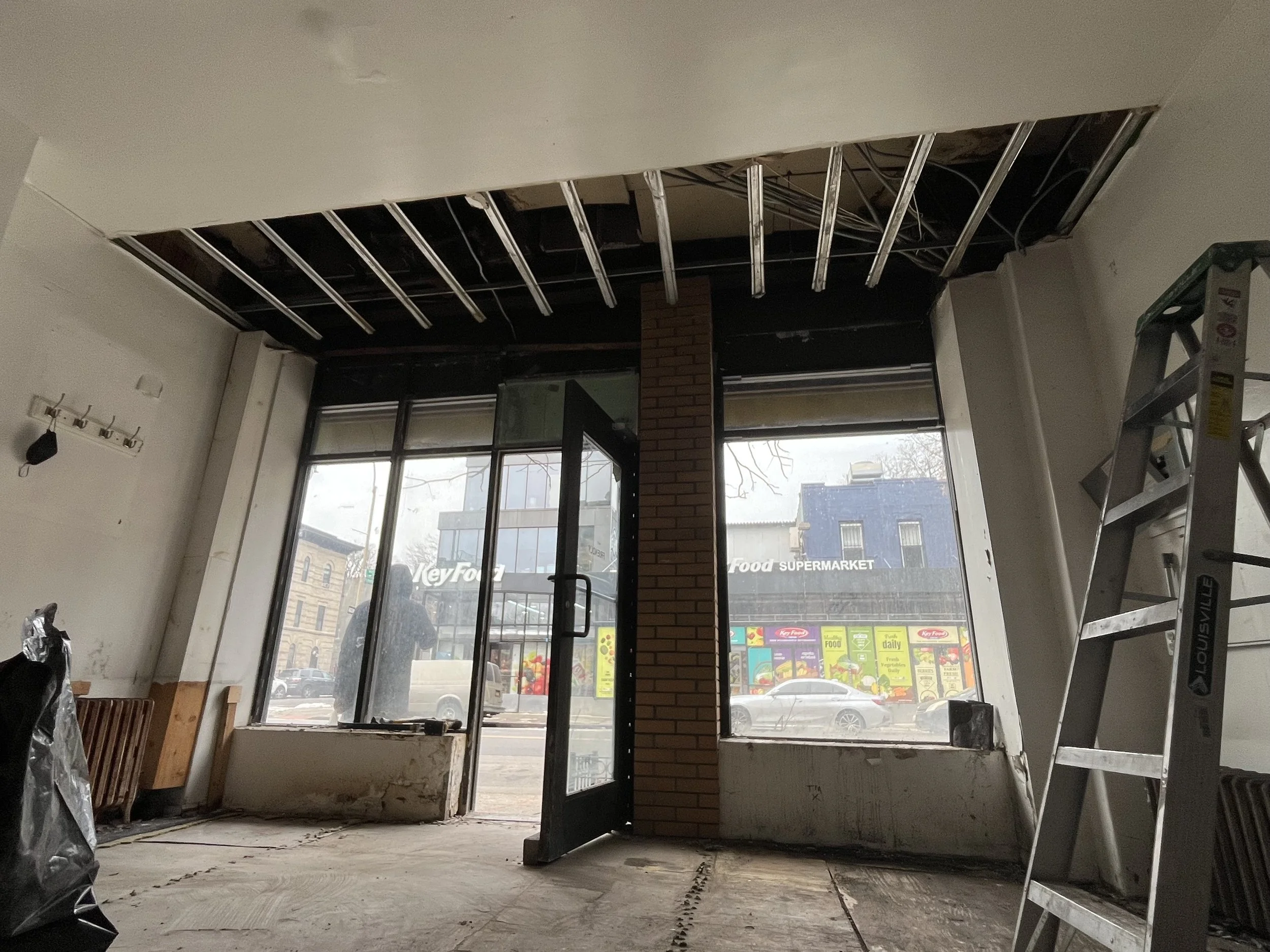 Interior of a building under renovation with exposed ceiling beams, ladder, and large front windows showing a street view.