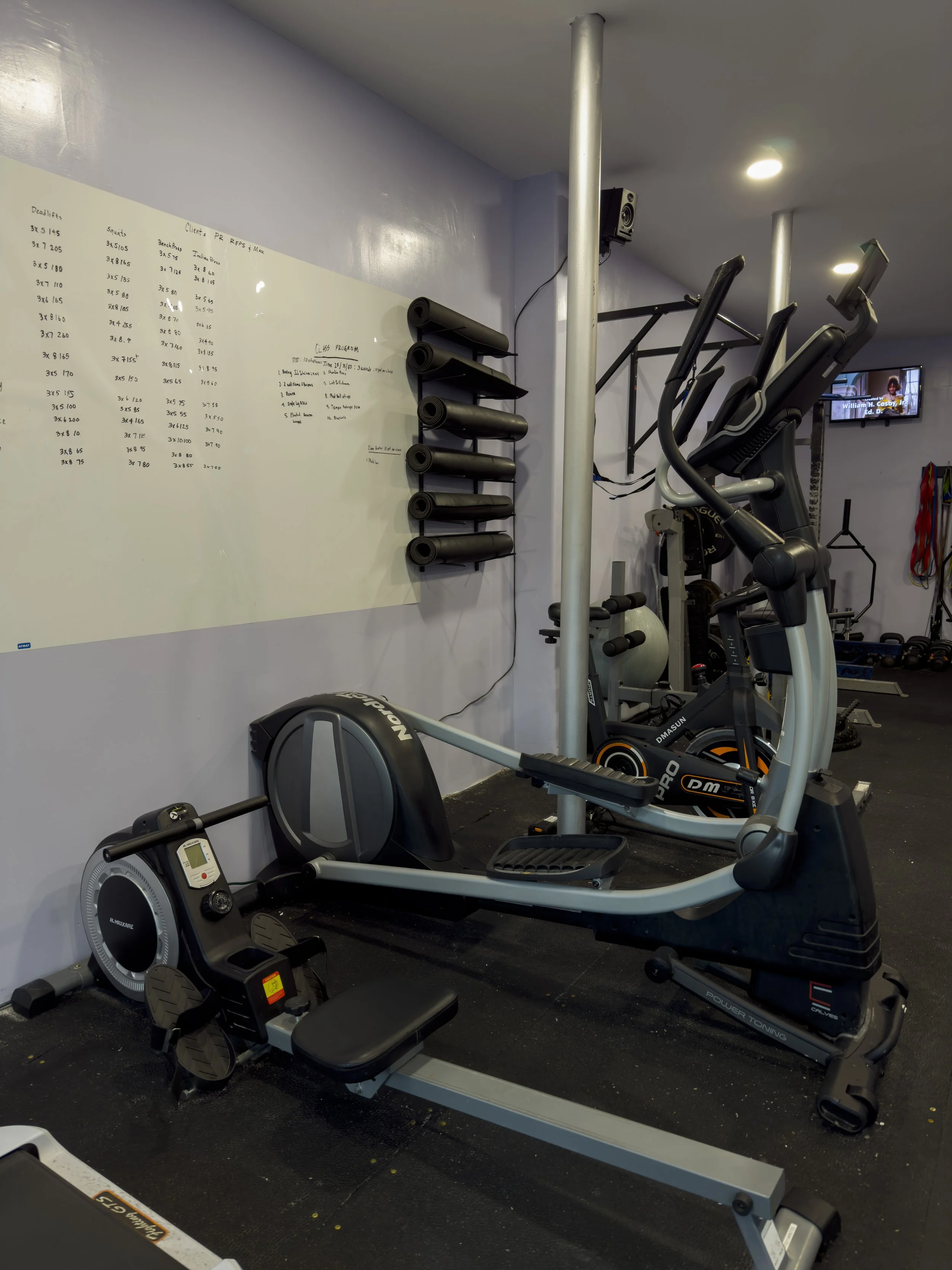 A gym with various exercise equipment, including a recumbent bike, elliptical machine, and rowing machine. A whiteboard with workout routines and a small television in the background.
