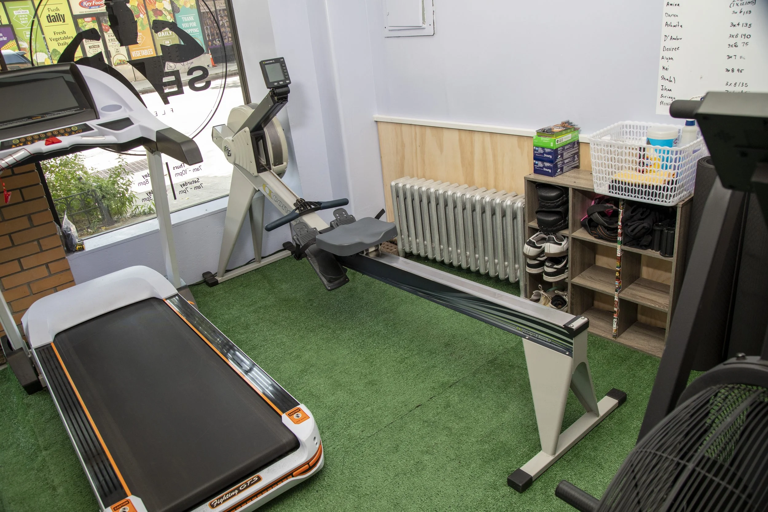 Indoor gym with a treadmill, exercise bike, and other fitness equipment on green carpet, a shelving unit with shoes and supplies, and a large window with store signage outside.