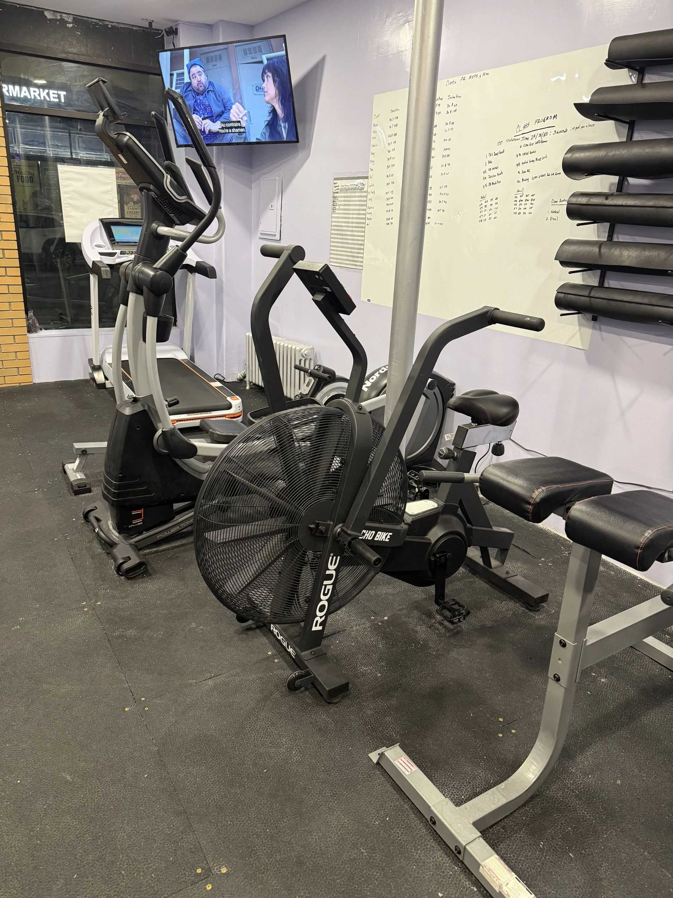 Interior of a gym with exercise equipment including a stationary bike, a treadmill, and a stair climber. A TV on the wall shows a program with two people talking. Whiteboards with workout schedules are on the wall, and some rolled mats are stacked on