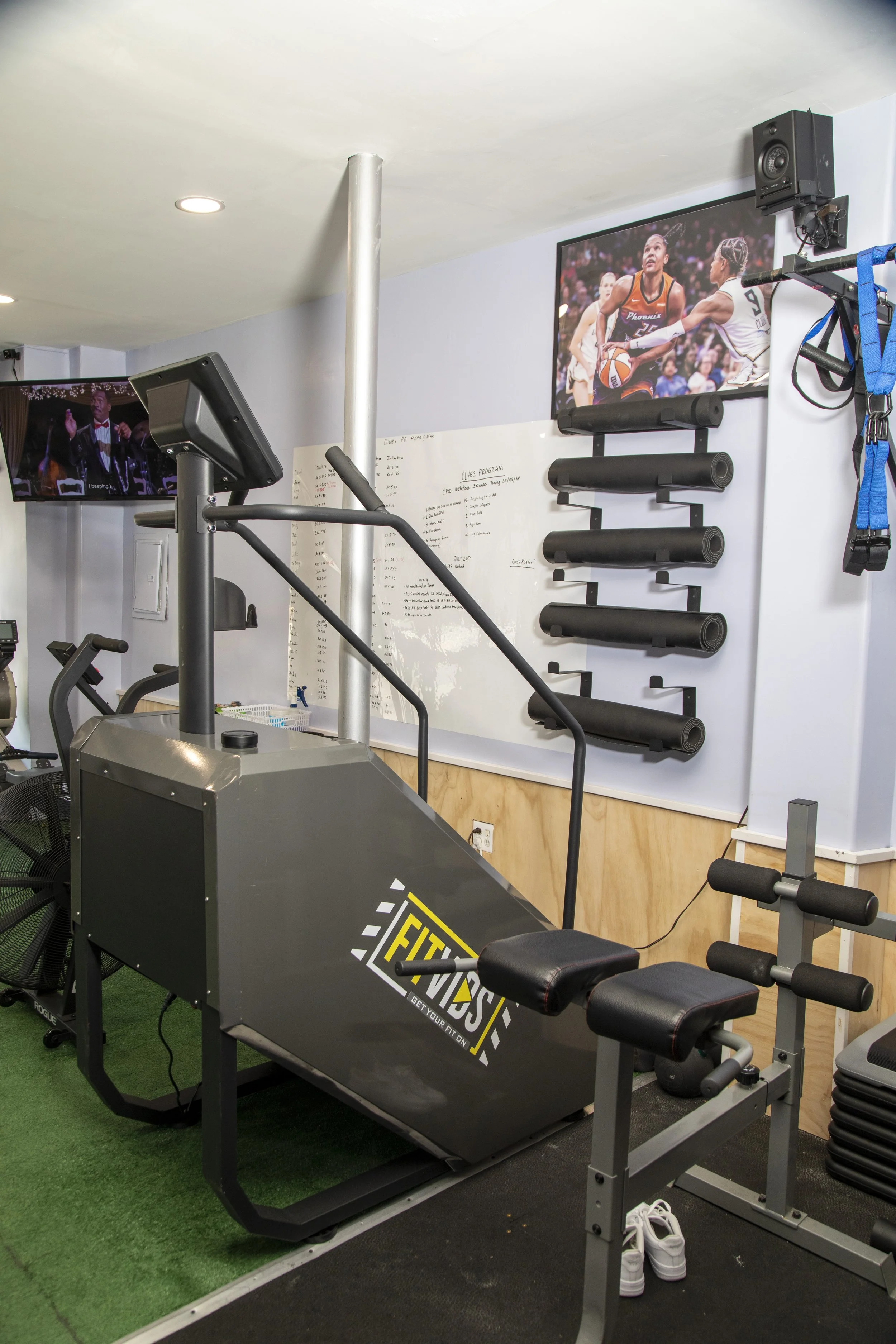 Gym with exercise equipment, including a black spray-painted treadmill with yellow and white lettering, foam rollers on the wall, and a TV showing a basketball game, with green and black rubber flooring.