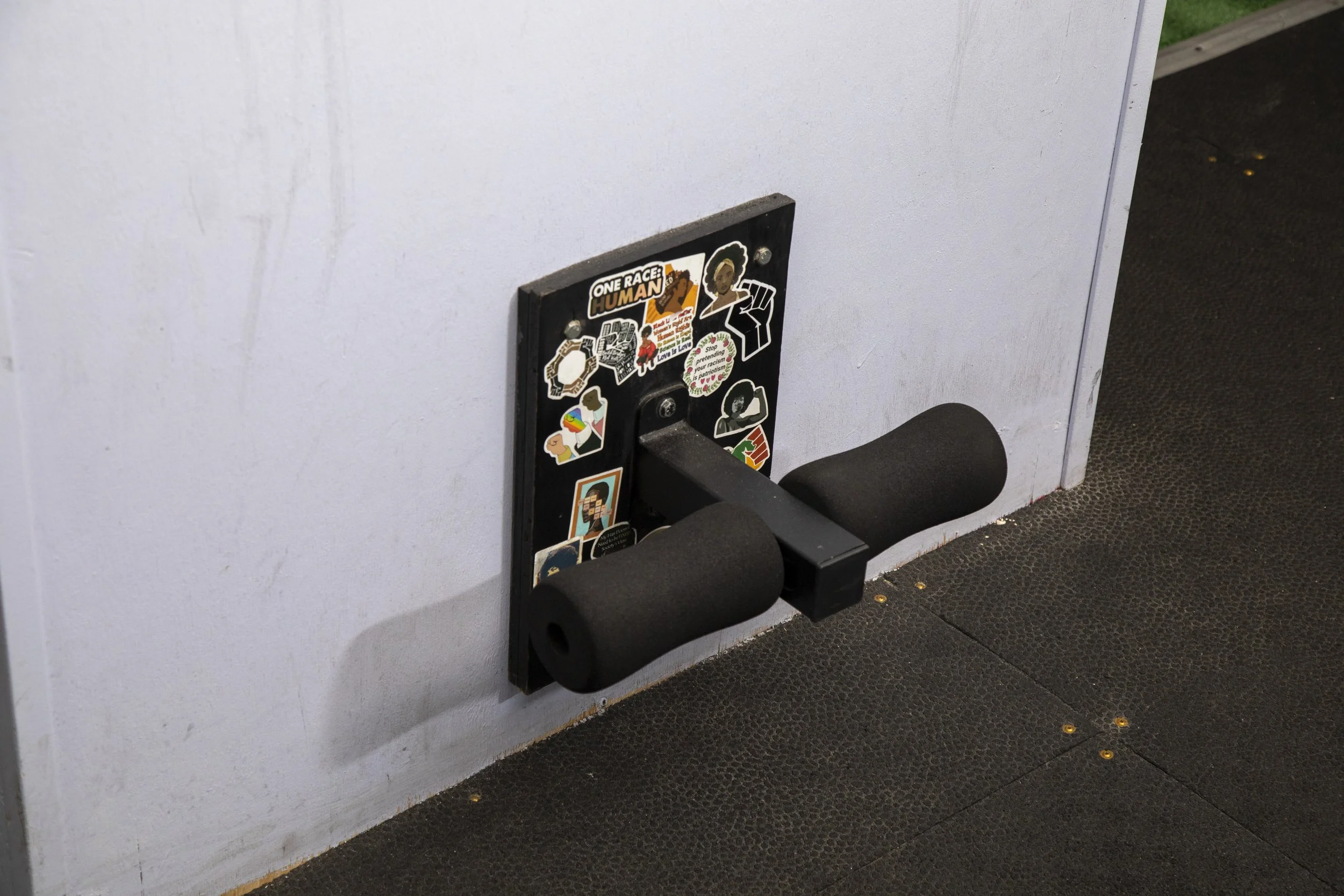 A black exercise step platform with a sticker that says 'One Race, Human' and various other stickers, positioned in front of a white wall and a black textured floor.