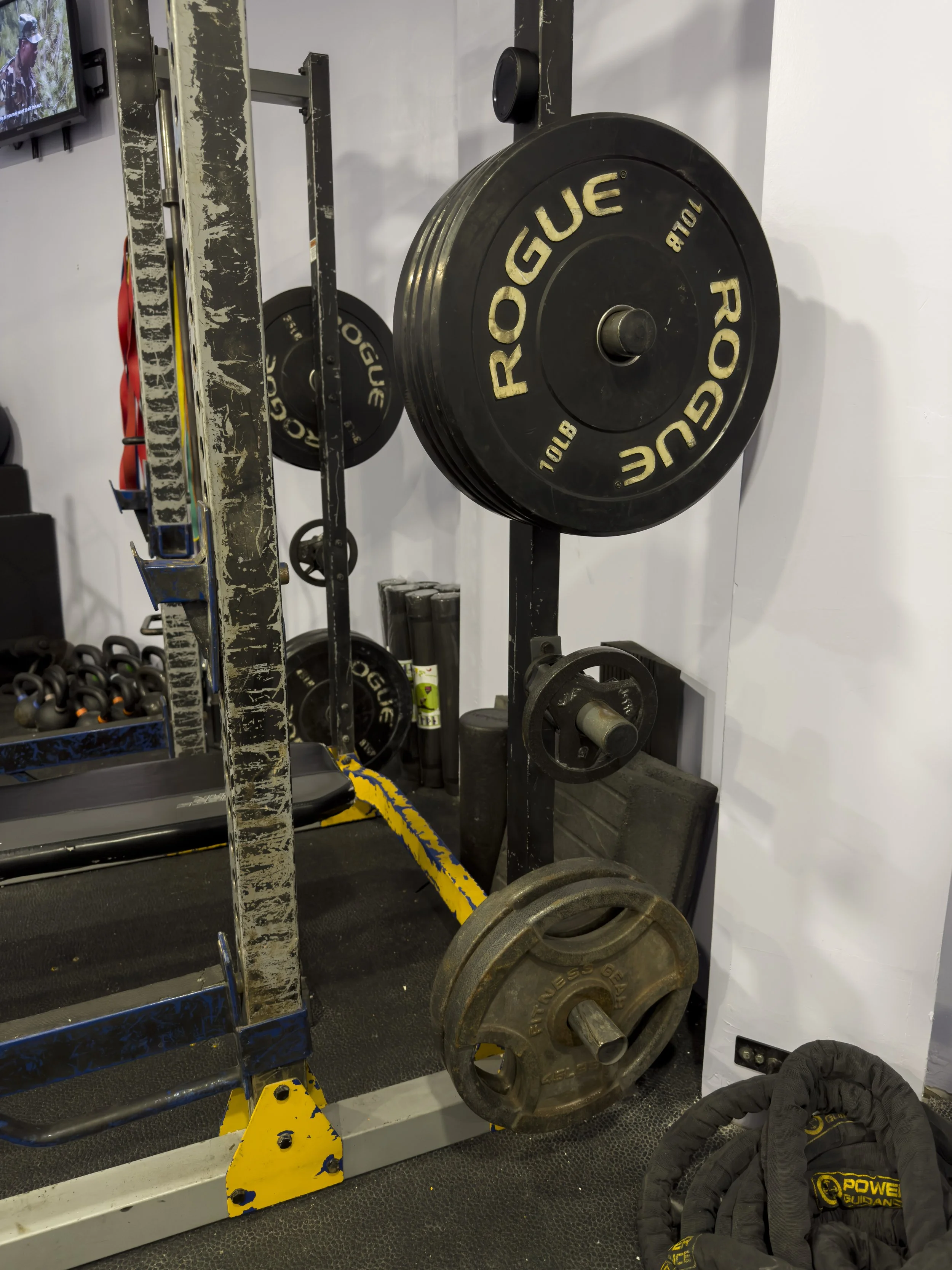 Weightlifting equipment in a gym, including black Rogue brand weight plates on a squat rack, with additional weight plates on the ground.