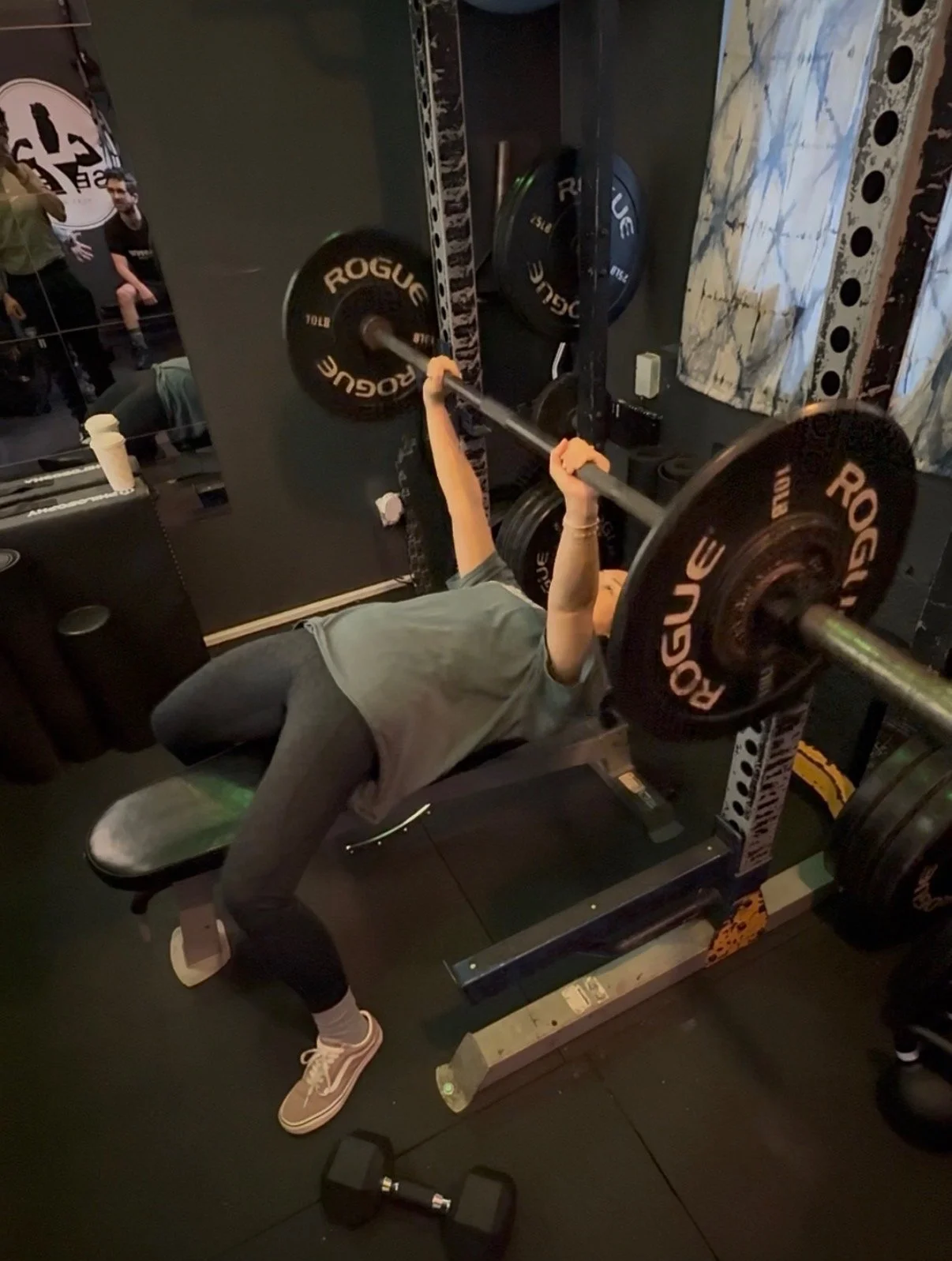 Person performing bench press exercise with barbell in a gym, with weight plates labeled 'Rogue' on the barbell.