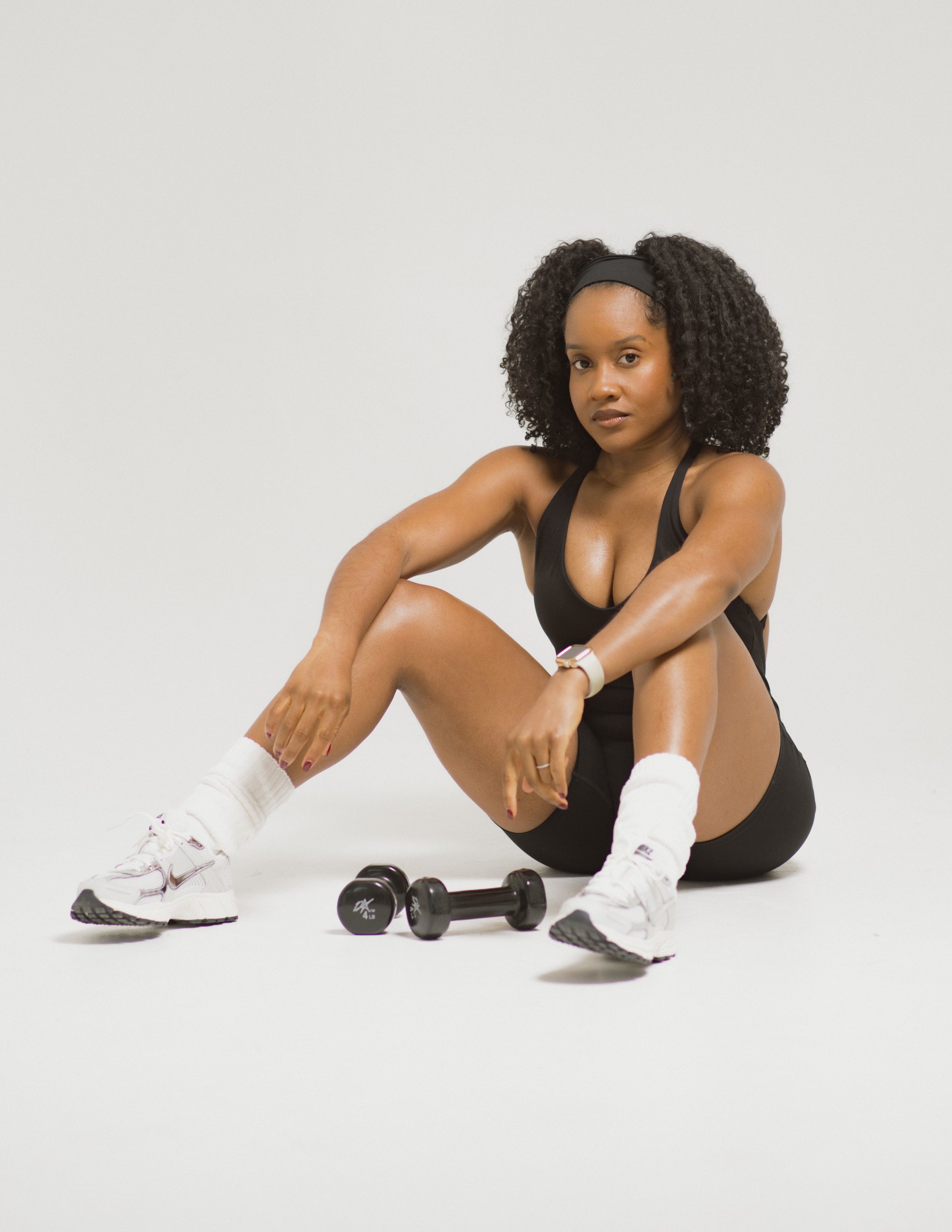 A woman sits on the floor wearing a black sports bra, black shorts, white sneakers, and a fitness tracker, with dumbbells in front of her, against a plain white background.