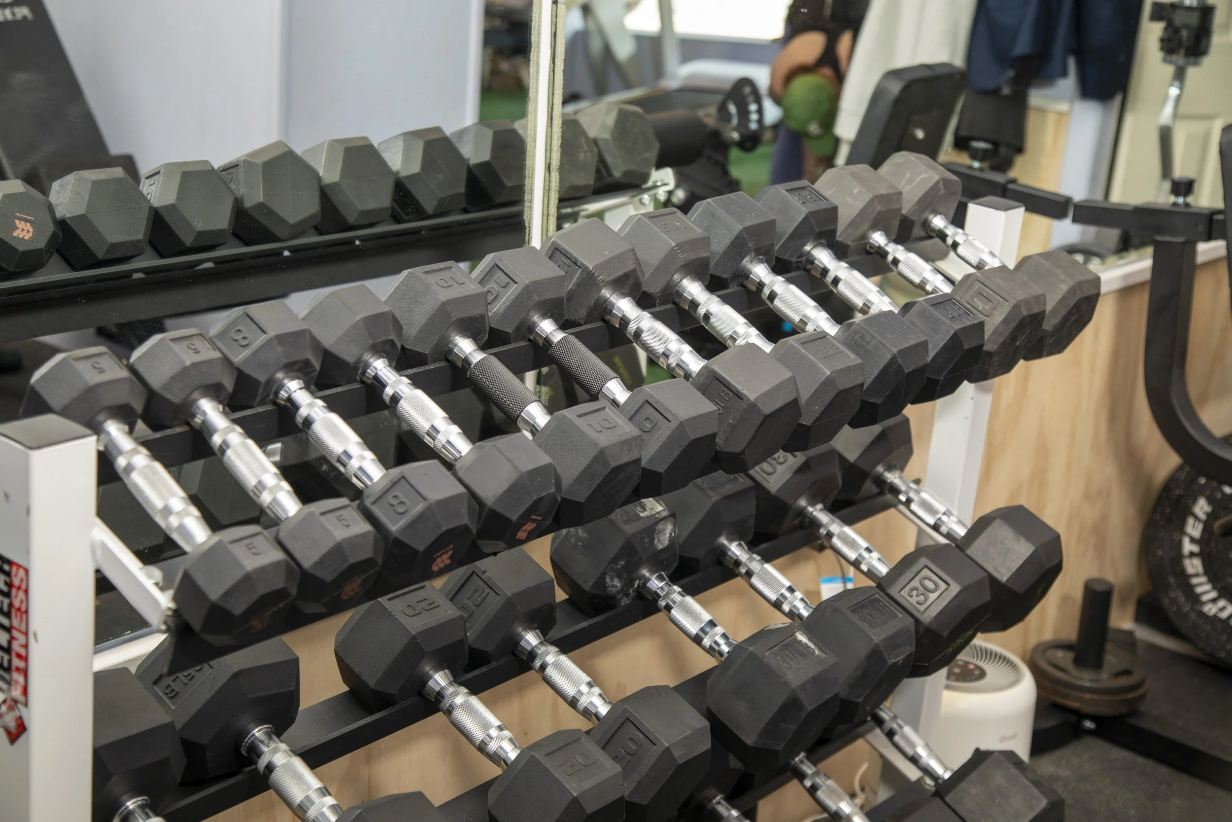 A rack of black hexagonal dumbbells with silver handles, arranged on multiple tiers in a gym setting. There is a mirror behind the rack reflecting some gym equipment and a person in the background.