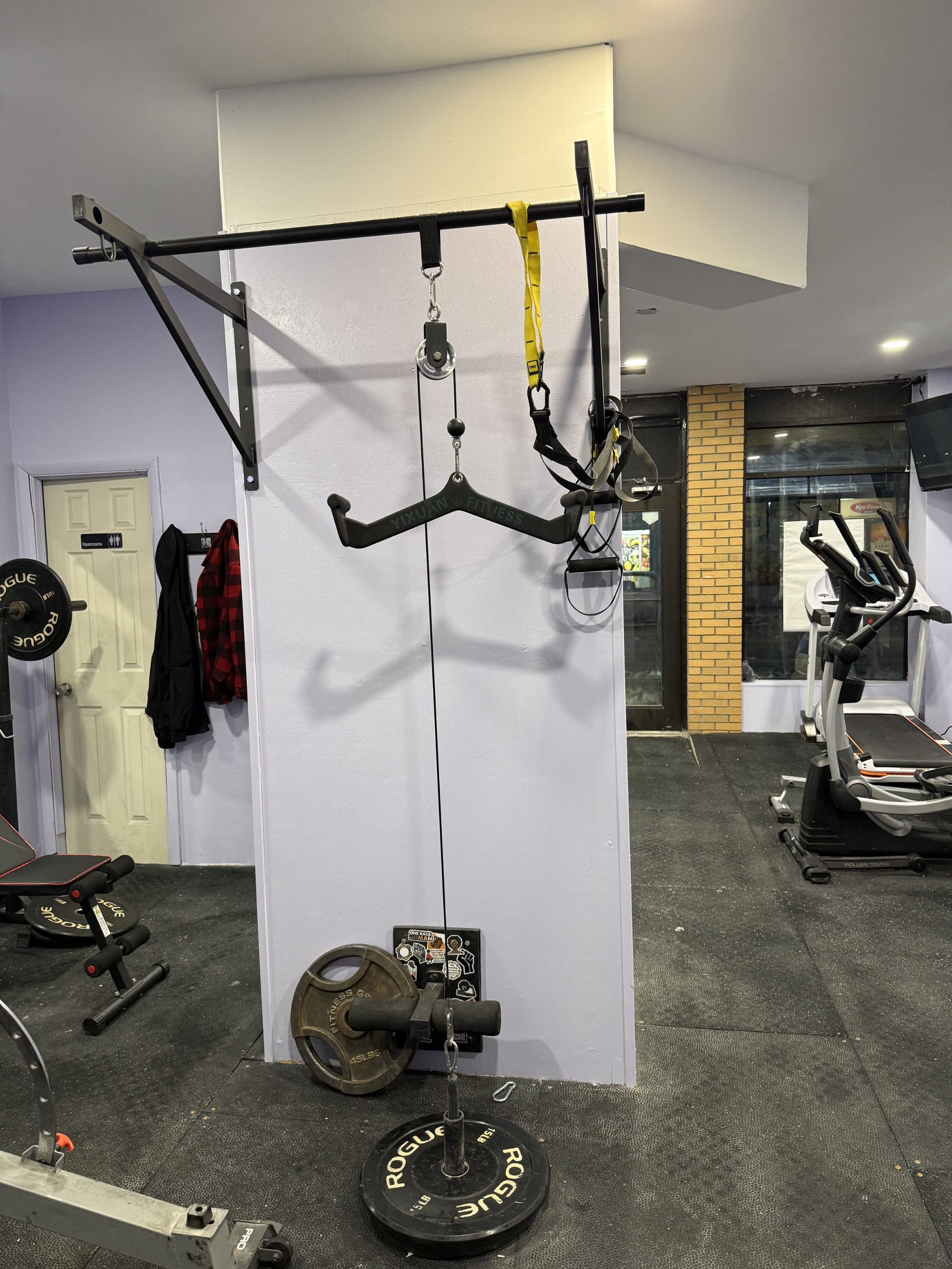 Gym gym with workout equipment, including a wall-mounted pull-up bar with resistance bands, a weight plate, and a treadmill in the background.