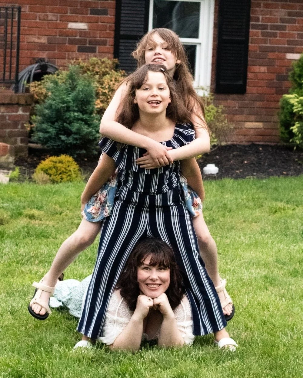 Three women forming a human pyramid on a lawn in front of a brick house, with shrubs and a window in the background.