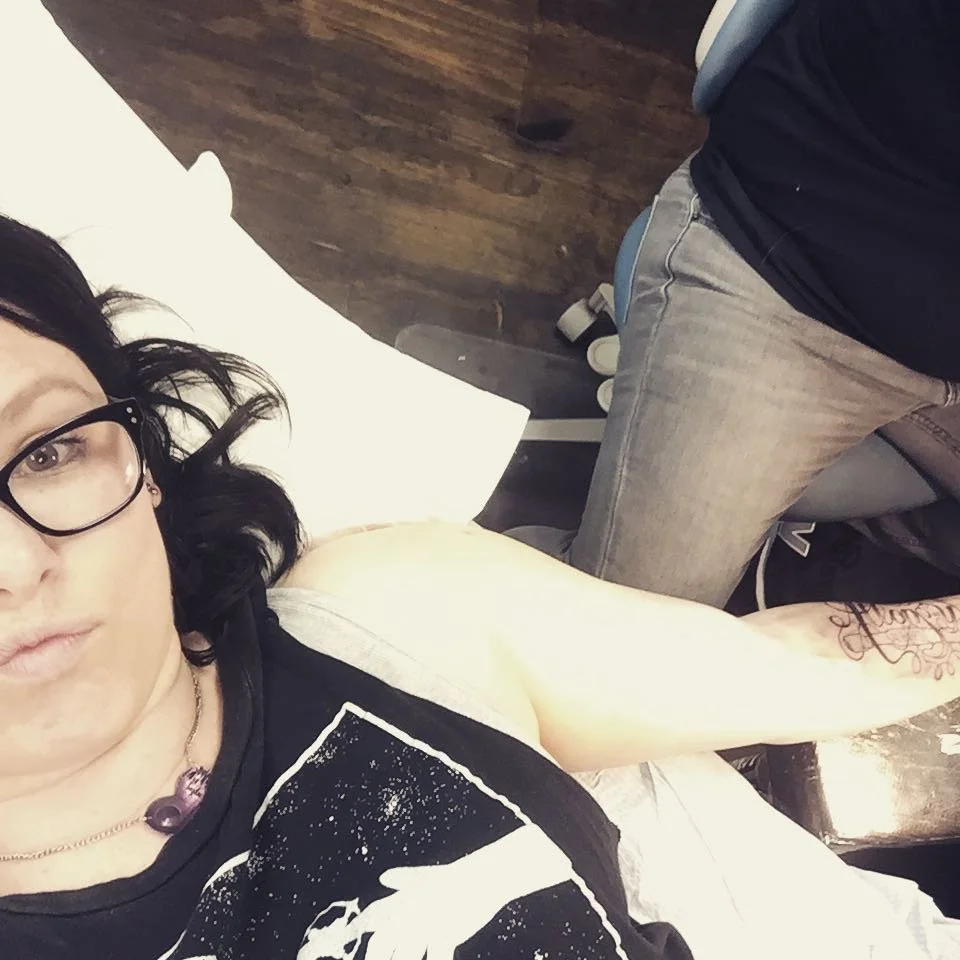 A woman with black hair, and glasses lying on a tattoo table, with a man sitting next to her, partially visible, tattooing her wearing gray jeans and a black shirt.