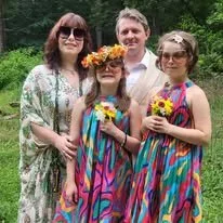 Four people, including a woman, a young girl, a teenage girl, and a teenage boy, standing outdoors with green trees in the background. The young girl and teenage girl are wearing colorful, striped dresses and holding sunflower bouquets, and the woman and teenage boy are casually dressed. The young girl is wearing a floral crown.