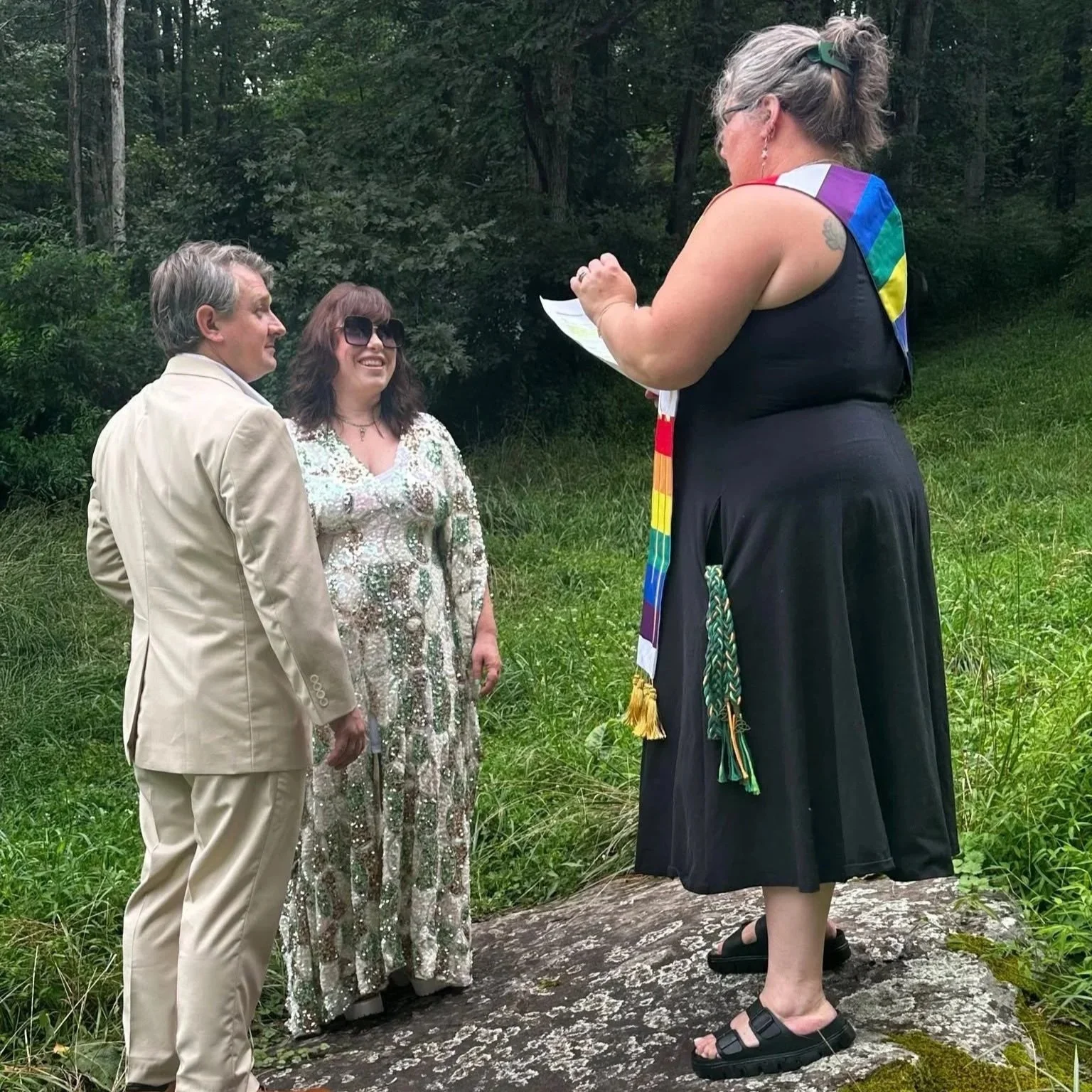 A small outdoor wedding ceremony with a couple standing on a large rock and a woman officiating, all surrounded by greenery. The couple is dressed in formal attire, and the officiant is wearing a black dress with a rainbow pride sash, short black sandals, and reading from a paper.