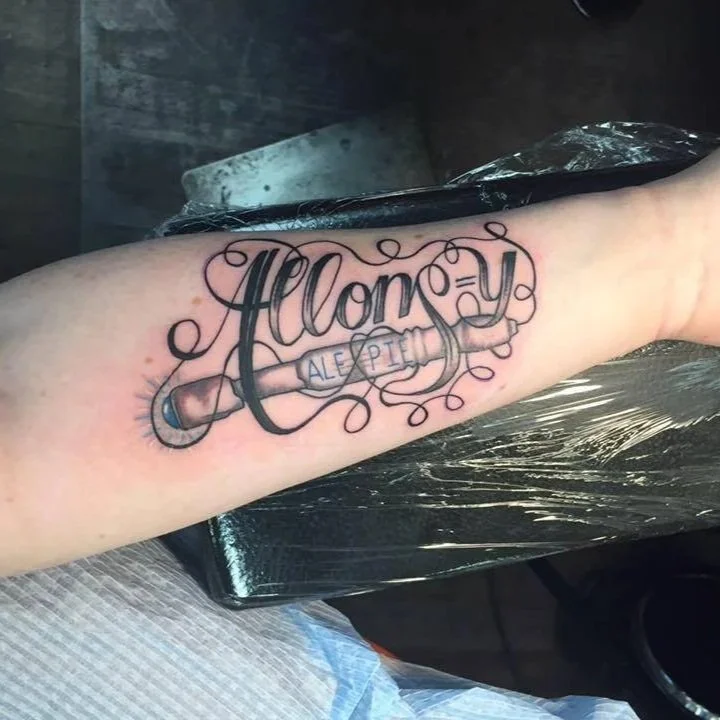 Tattoo on forearm with the word "Allory" in cursive, incorporating a syringe with the words "Alex Piem" on it, surrounded by decorative swirls.