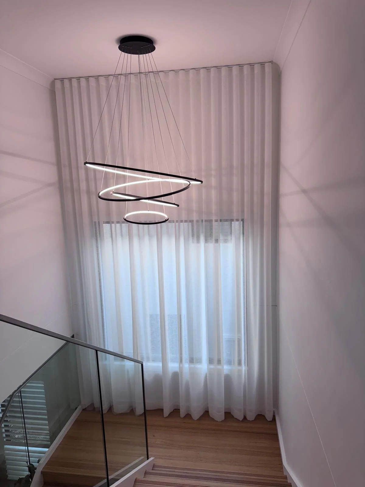 Modern spiral chandelier hanging from ceiling in minimalist home interior with tall white curtains and a glass railing overlooking a staircase.