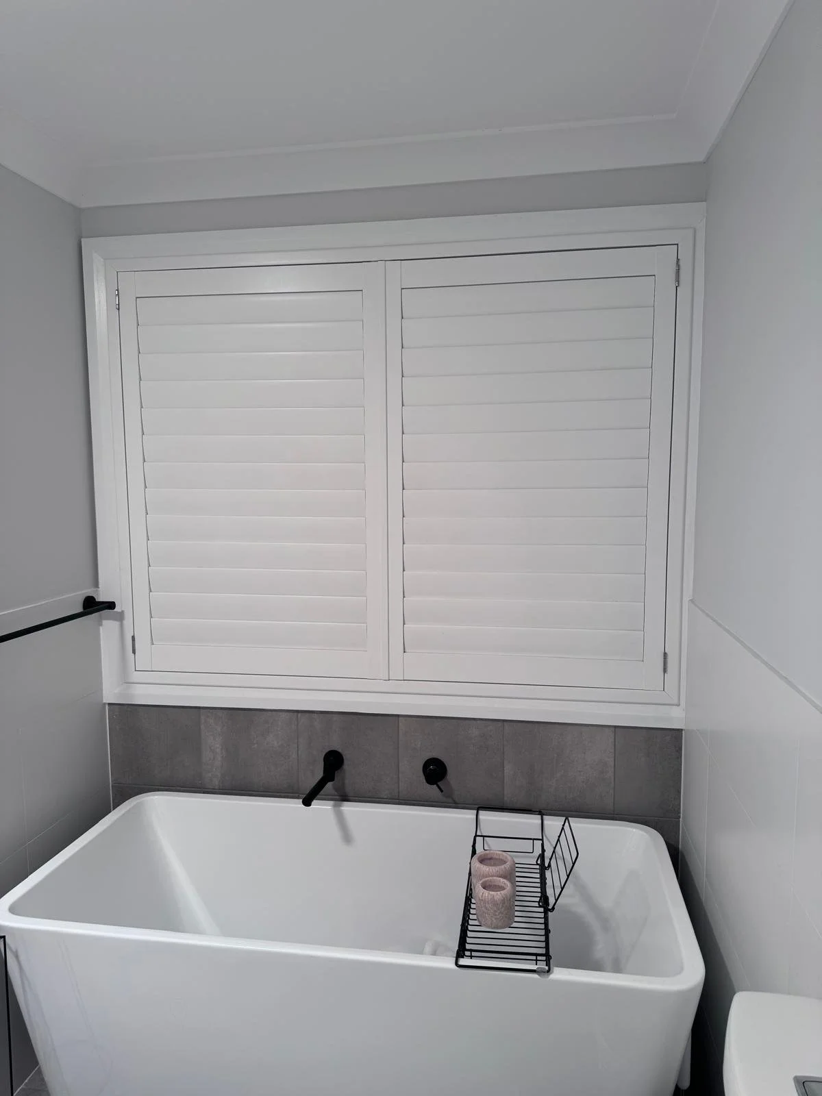 A white bathtub with black fixtures, a wire basket holding two rolls of toilet paper, grey tiled wall behind it, and white shutters on the window.