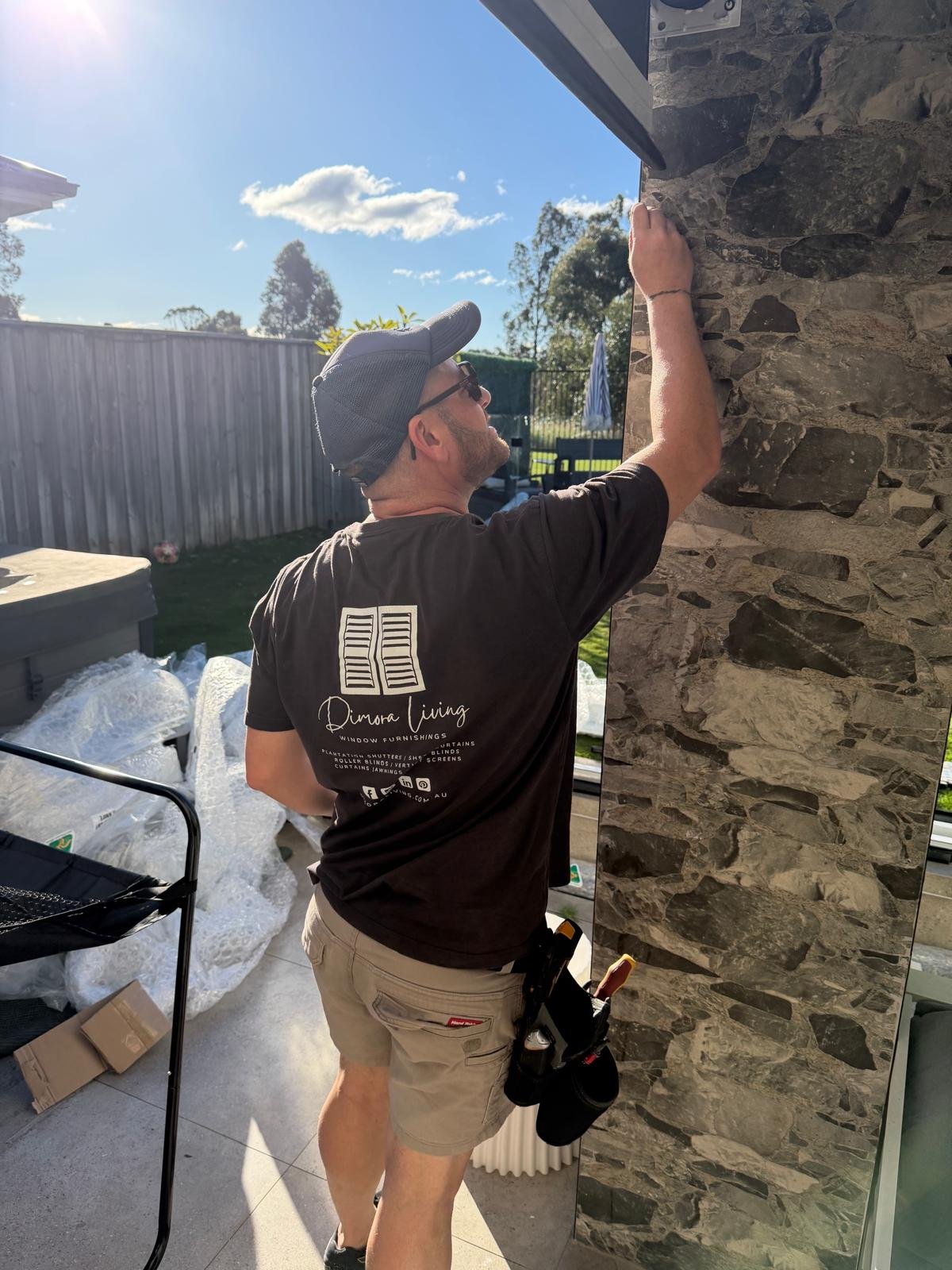 A man wearing a gray cap, sunglasses, and a black T-shirt is installing or working on a stone or wood panel on a wall outside. The back of his T-shirt reads "Dimora Living" with a logo and list of window furnishing services. He is holding a tool in his right hand and has a black pouch or bag attached to his belt. The background shows a sunny backyard with a fence, some trees, and outdoor furniture.