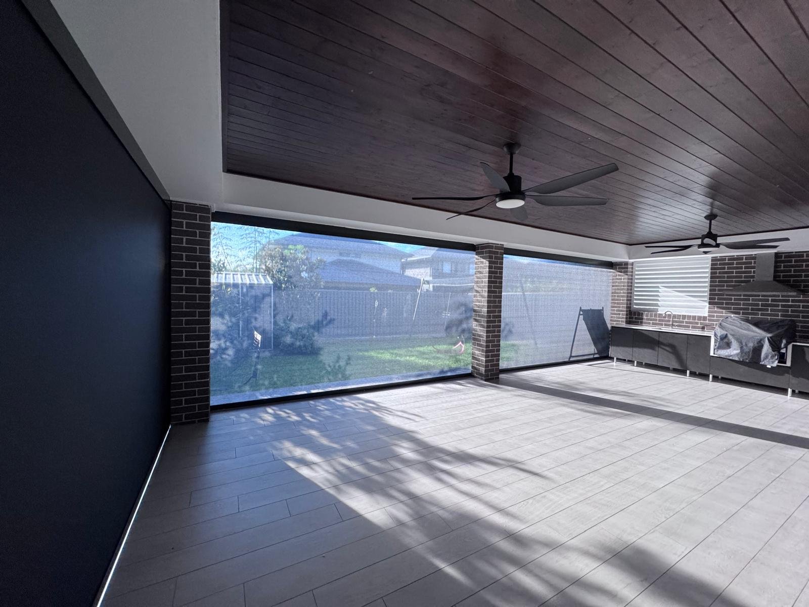 Bright indoor patio with wooden ceiling, ceiling fans, brick support columns, large sliding screen doors covering the backyard, and a black outdoor kitchen with a grill covered in plastic, with shadows cast on the light-colored tile floor.