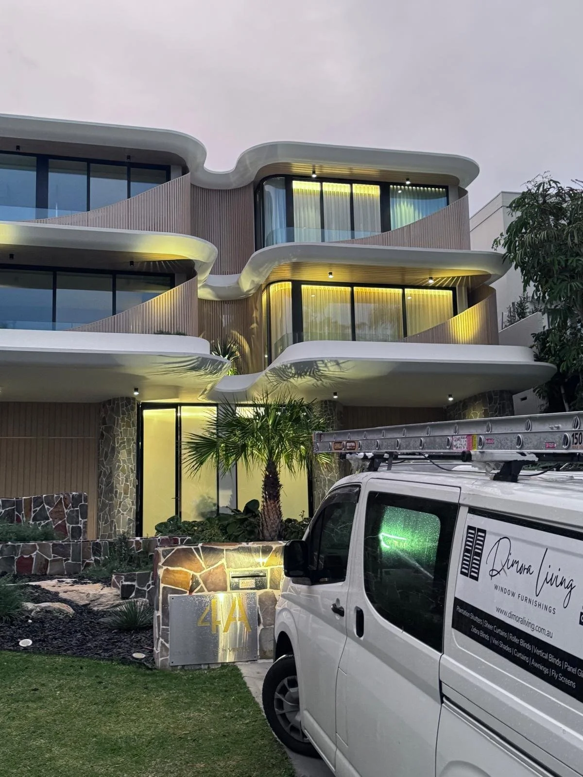Modern multi-story building with rounded balconies illuminated by yellow lights, glass walls, and tropical plants in front. A white van with company branding is parked outside.