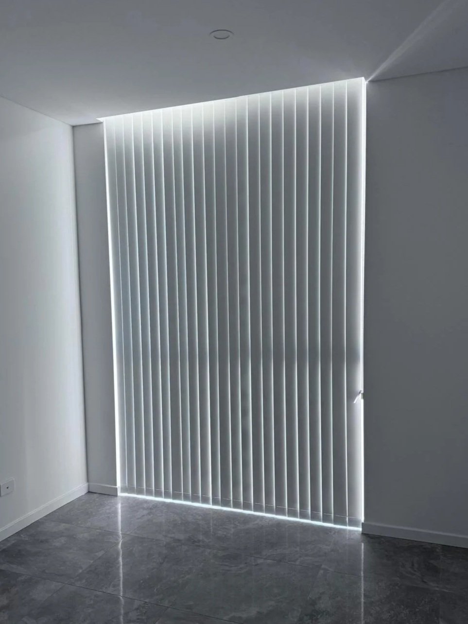 Modern interior corner of a room with vertical white blinds partially open, allowing light to shine through.