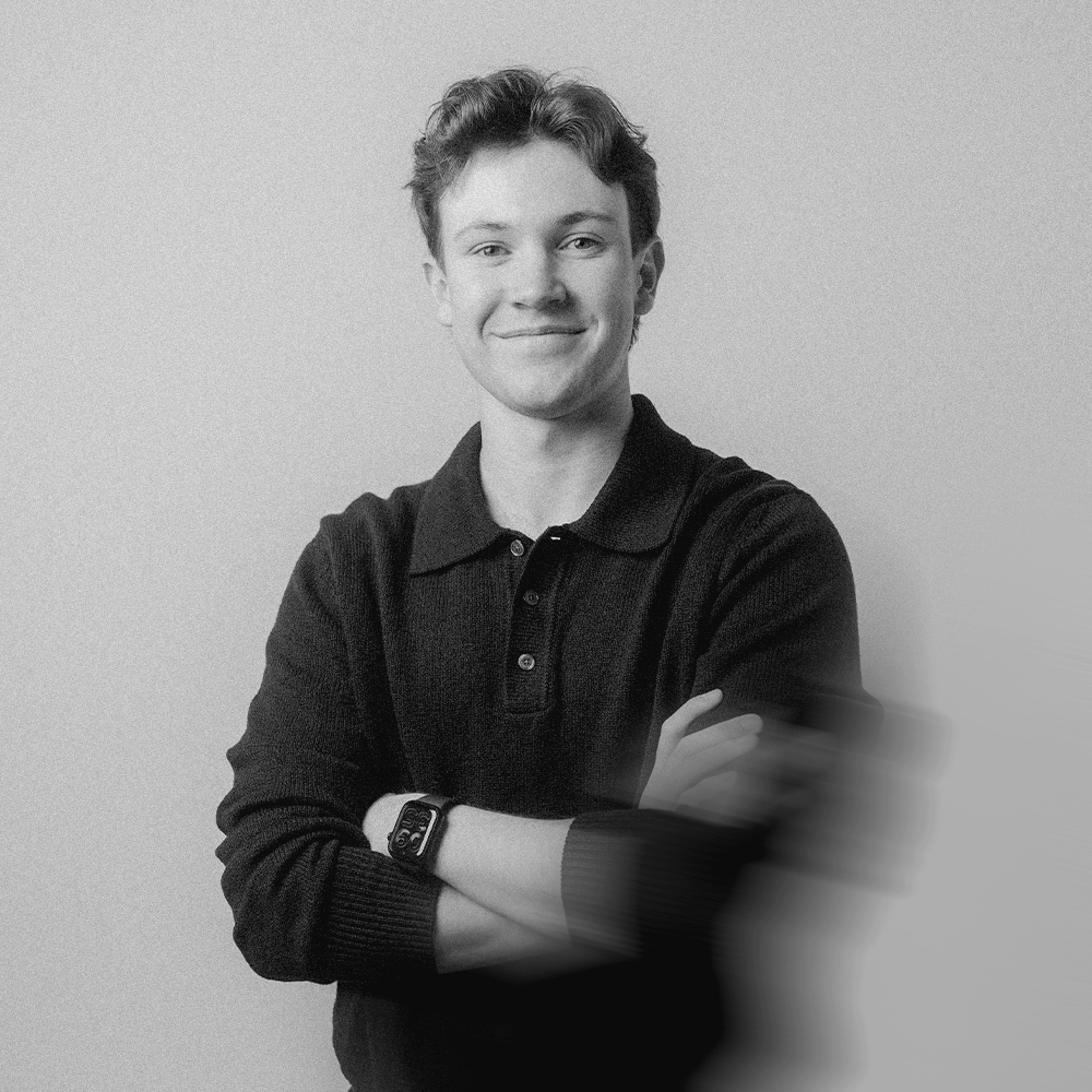 Black and white photo of a young man with short, wavy hair, smiling, wearing a dark long-sleeve polo shirt with folded arms, standing against a plain background. Slight motion blur on his right arm.