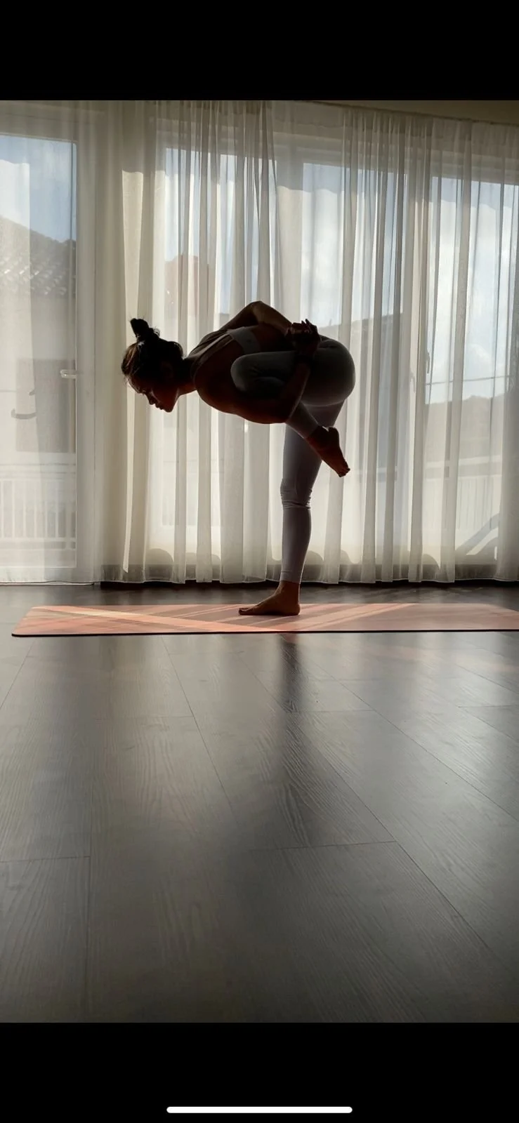 A woman practicing yoga indoors, balancing on one leg while holding her other leg behind her back, with sheer curtains and sunlight in the background.