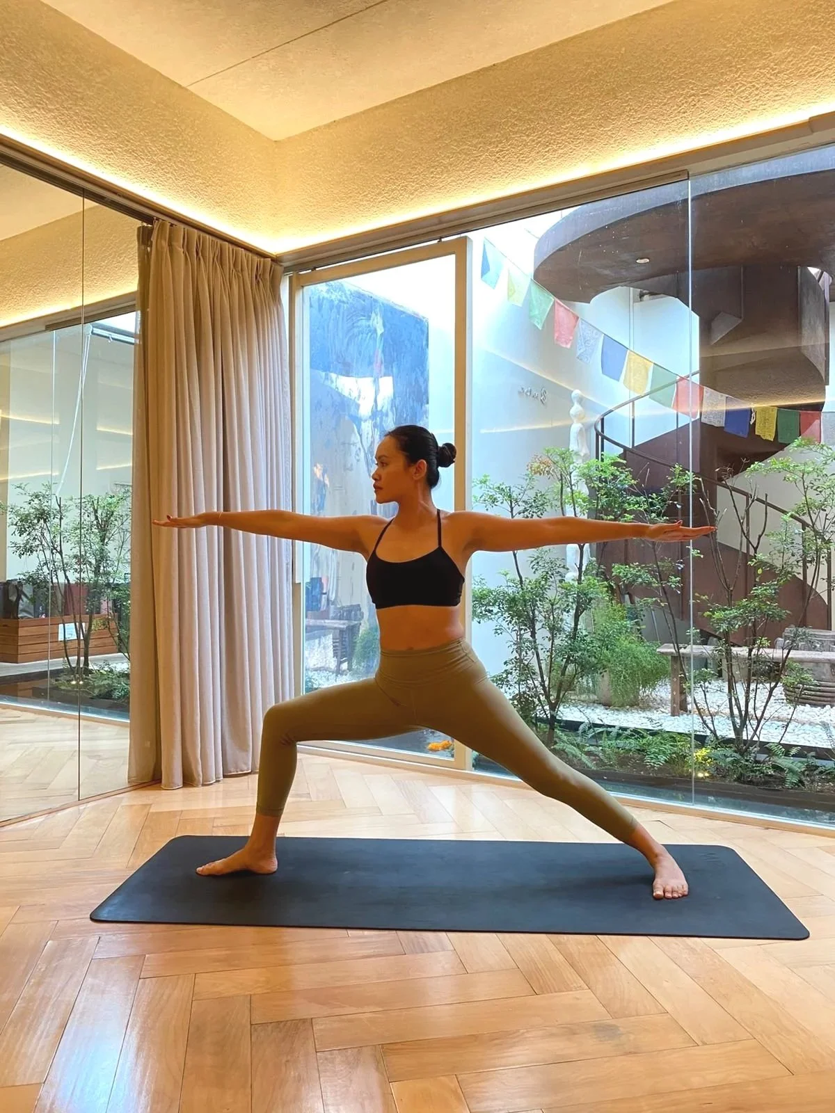 A woman practicing yoga indoors, balancing on one leg while holding her other leg behind her back, with sheer curtains and sunlight in the background.