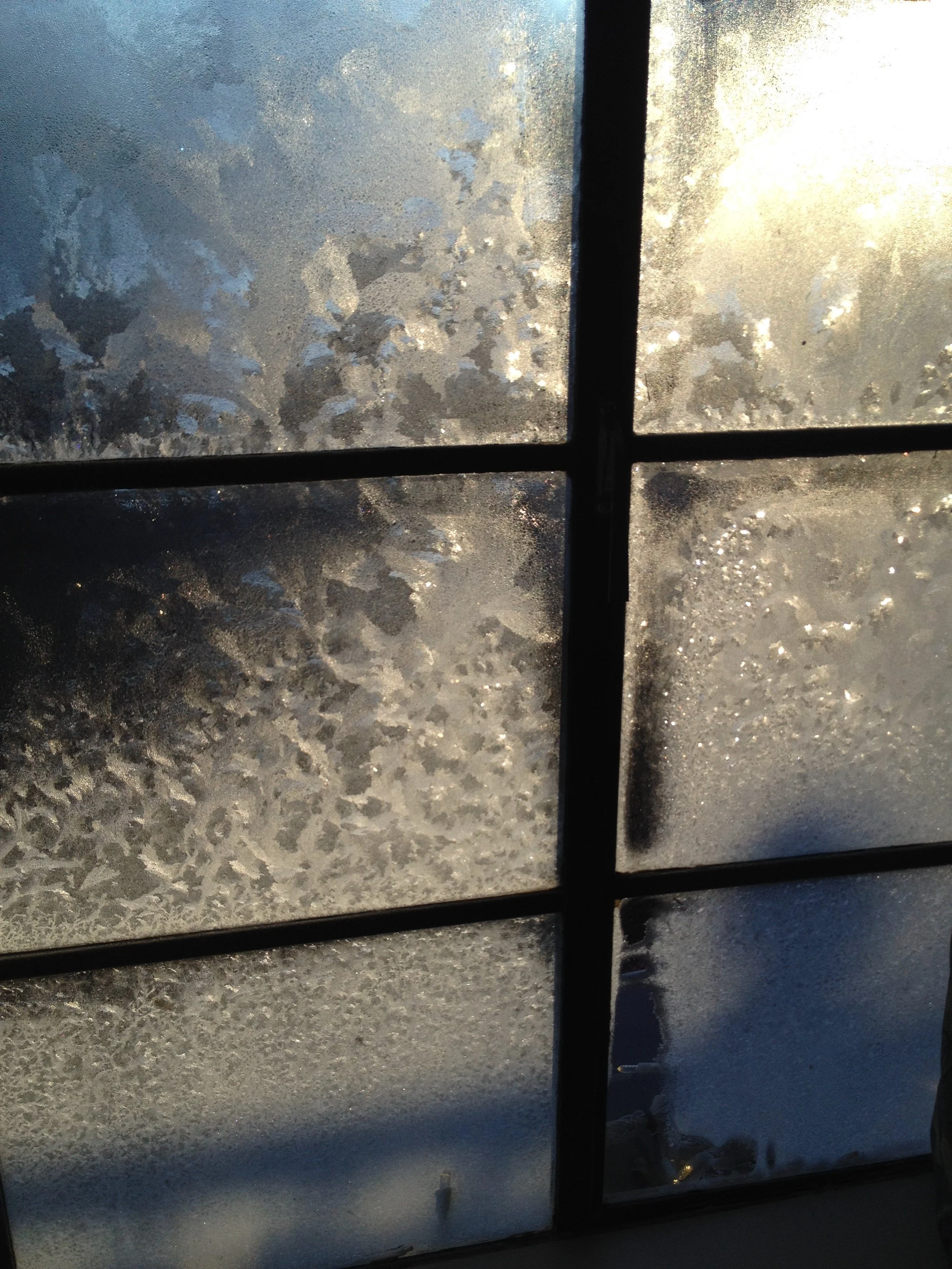 Frosted window panes with snow and ice accumulation, divided by black window frames, with sunlight shining through.