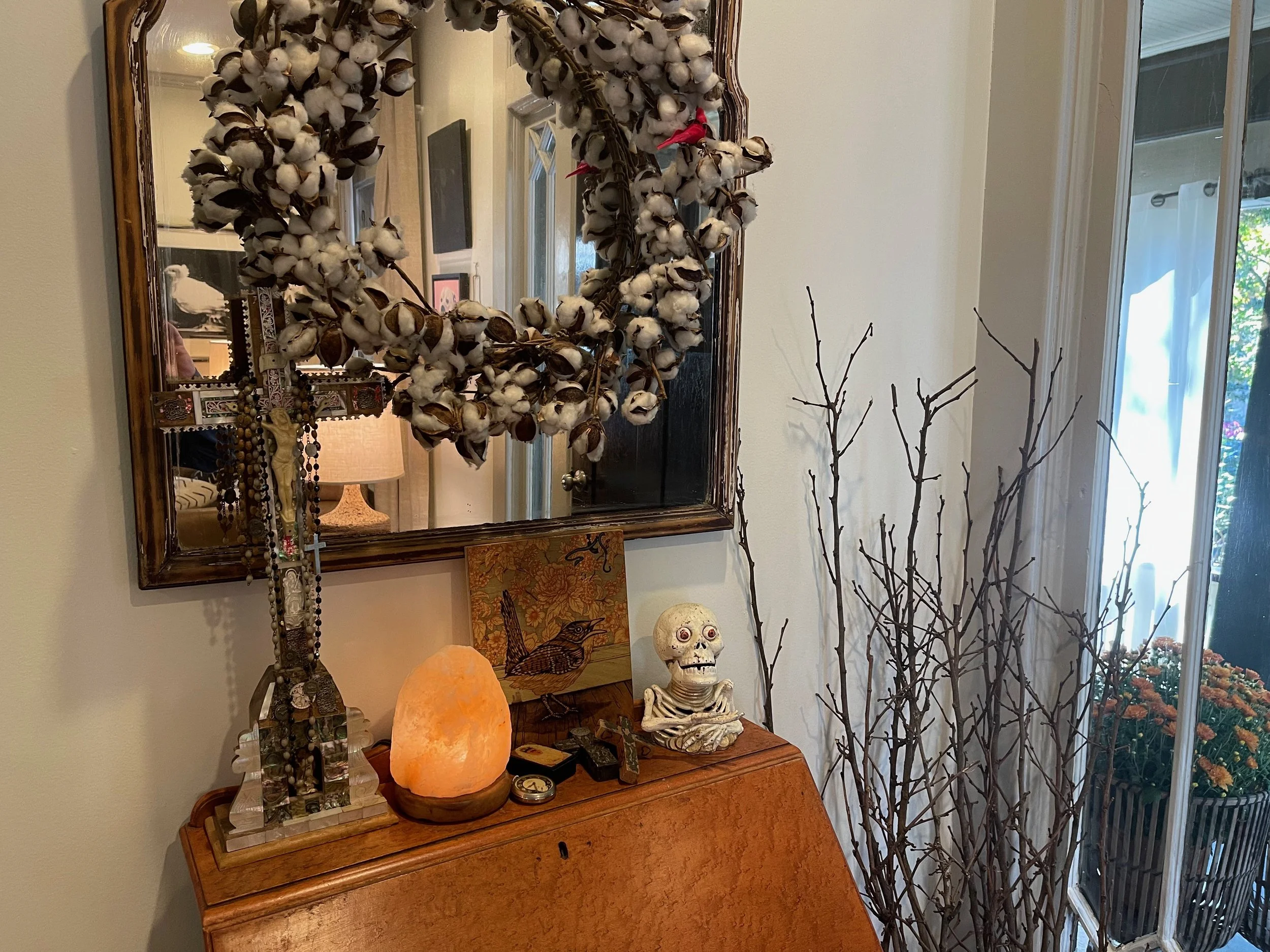 Decorative home entryway with mirror, cotton boll wreath, jewelry, salt lamp, skeleton figurine, and bare branches near window.