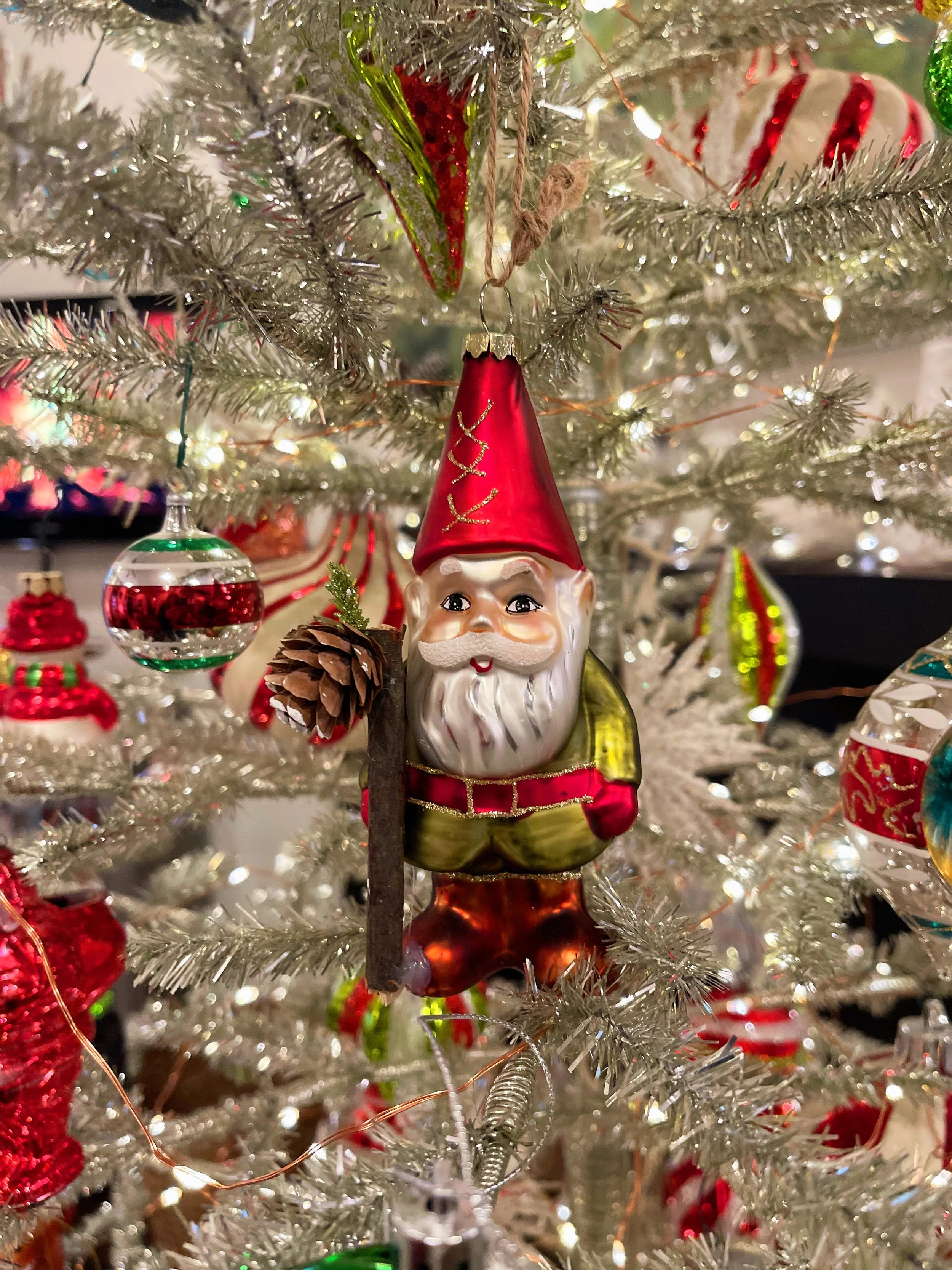 Decorative Christmas tree ornament of a gnome with a white beard, red hat, green and red clothes, holding a pinecone, hanging on a decorated Christmas tree.