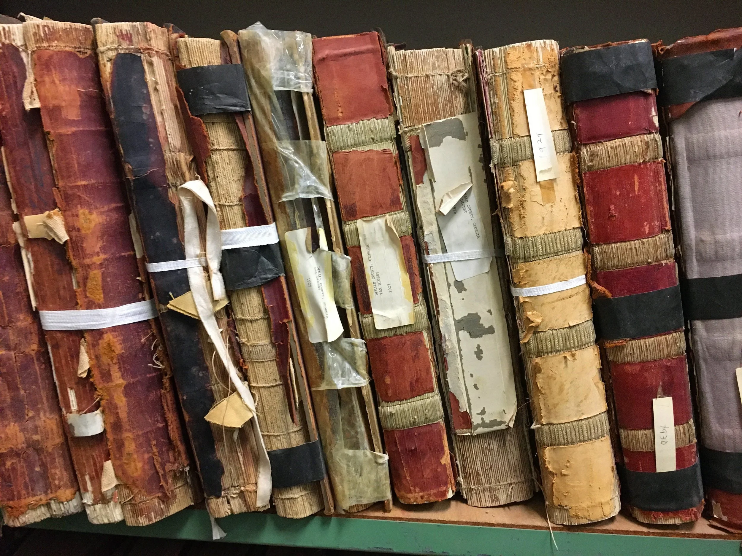 Row of old, worn books with frayed, discolored covers, some with labels and tape, stacked on a surface.