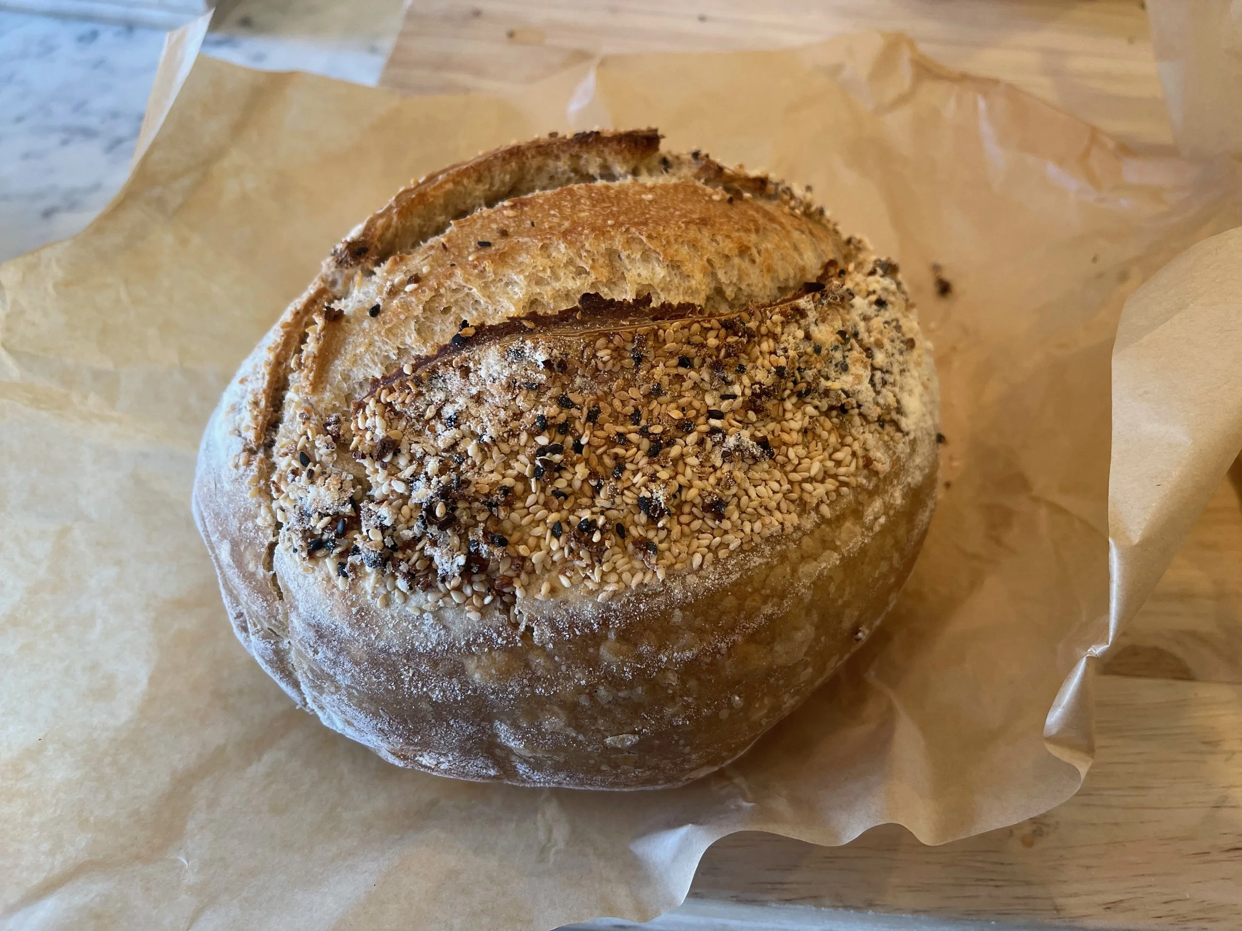A bread roll topped with sesame seeds and black pepper, placed on crumpled parchment paper on a wooden surface.
